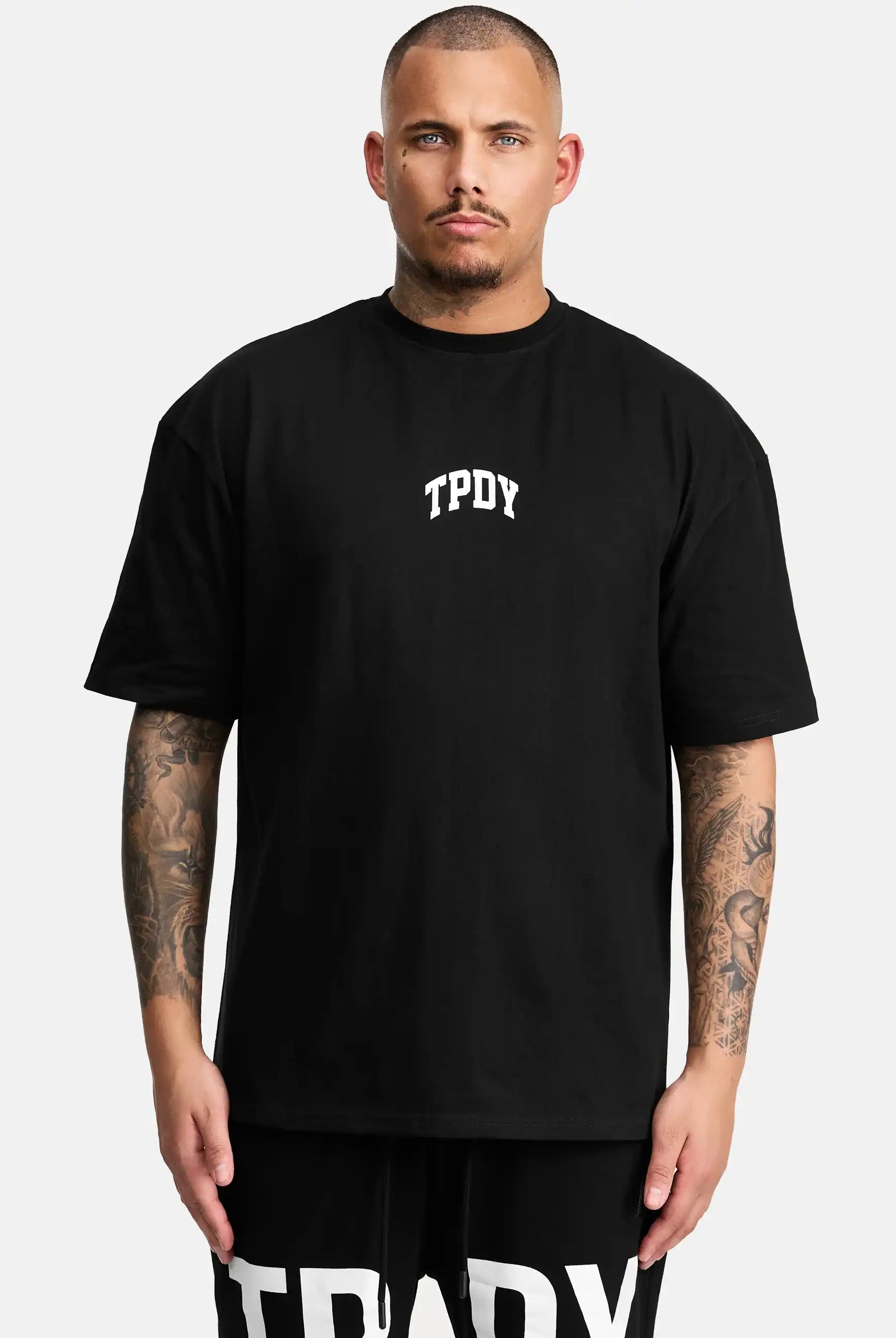Ein Mann mit kurzgeschnittenen Haaren und tätowierten Armen modelliert das trueprodigy Elijah Oversized T-Shirt mit Druck. Es hat eine lockere Passform und einen auffälligen weißen TPDY-Print auf der Brust und steht vor einem schlichten hellen Hintergrund.