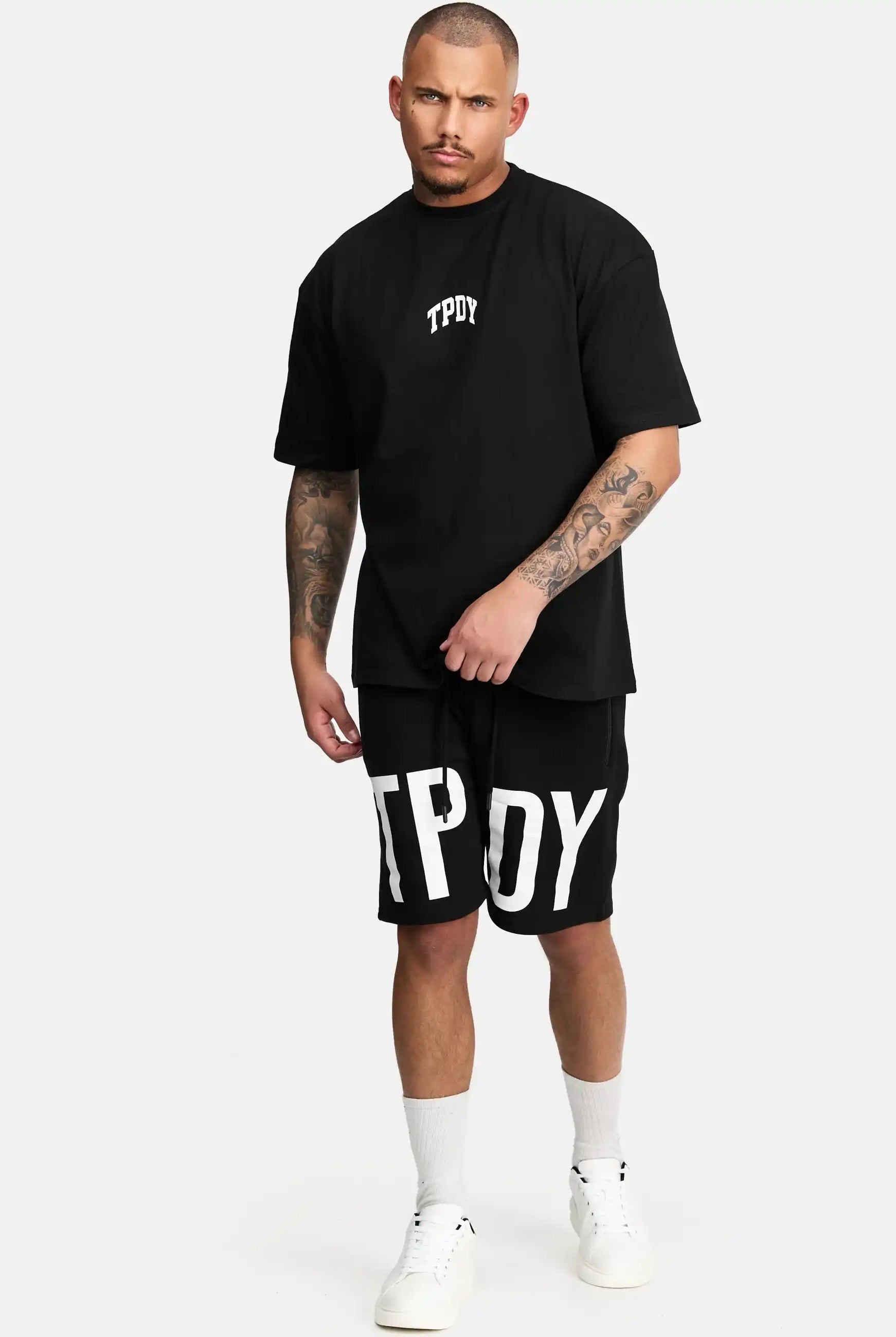 Ein Mann mit kurzen Haaren und tätowierten Armen trägt das schwarze trueprodigy Elijah Oversized T-Shirt mit Druck und passende Shorts, beides mit weißem TPOY-Aufdruck, sowie weiße Socken und Turnschuhe vor einem schlichten hellen Hintergrund.