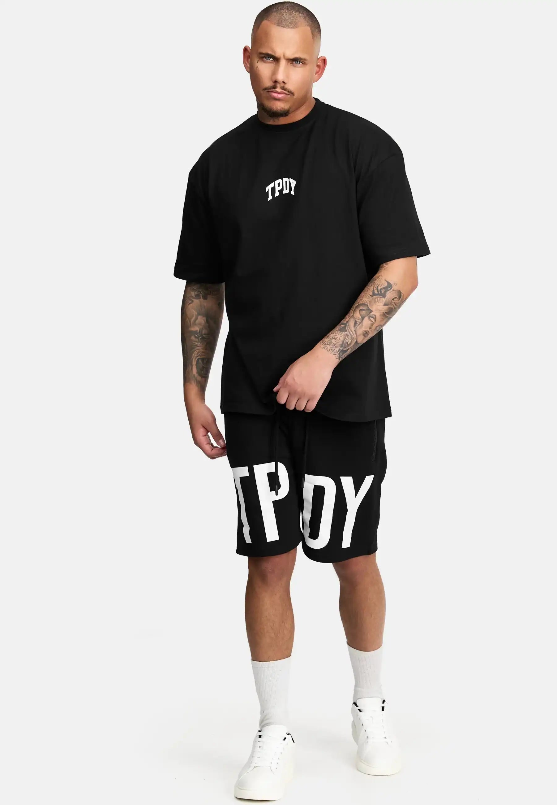 Ein Mann mit kurzen Haaren und tätowierten Armen trägt das schwarze trueprodigy Elijah Oversized T-Shirt mit Druck und passende Shorts, beides mit weißem TPOY-Aufdruck, sowie weiße Socken und Turnschuhe vor einem schlichten hellen Hintergrund.