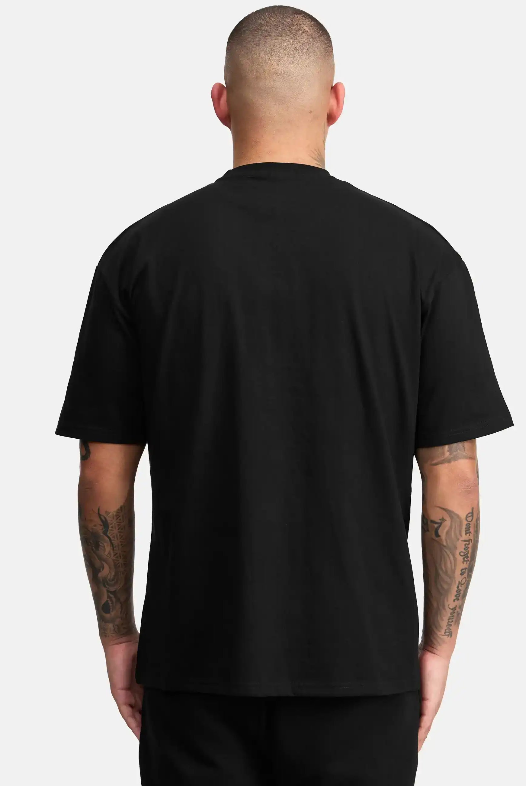 Eine Person mit kurzen Haaren ist von hinten zu sehen, die das trueprodigy Channing Oversized T-Shirt mit Druck trägt, gepaart mit einer schwarzen Hose. Beide tätowierten Arme sind sichtbar, und der schwere Jersey-Stoff hebt sich von einem hellgrauen Hintergrund ab.