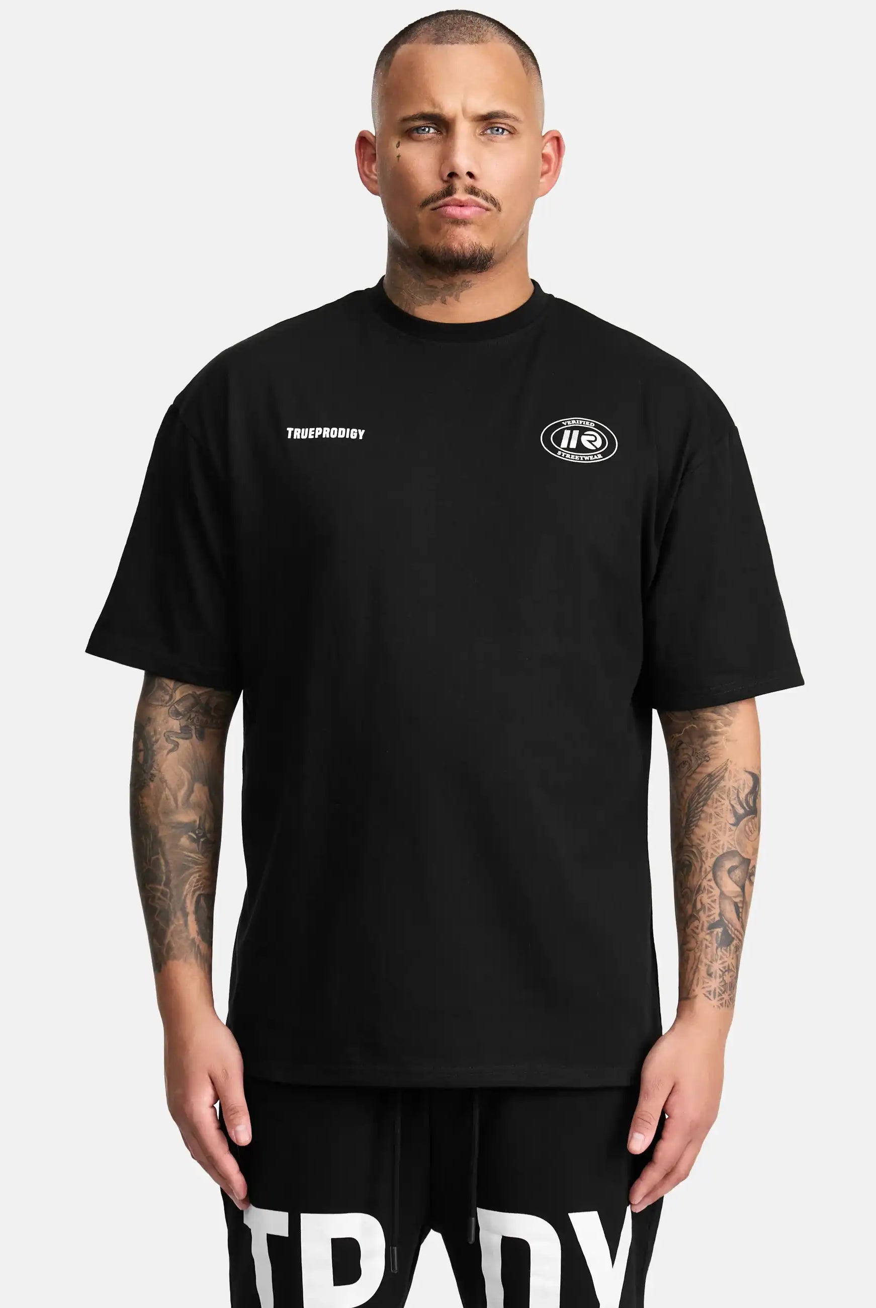 Ein Mann mit kurzen Haaren und Tattoos trägt das Channing Oversized T-Shirt mit Druck von trueprodigy - ein übergroßes Unisex-T-Shirt mit kleinen weißen Grafiken auf der Brust - und schwarze Shorts mit fetten weißen Schriftzügen vor einem schlichten hellen Hintergrund.