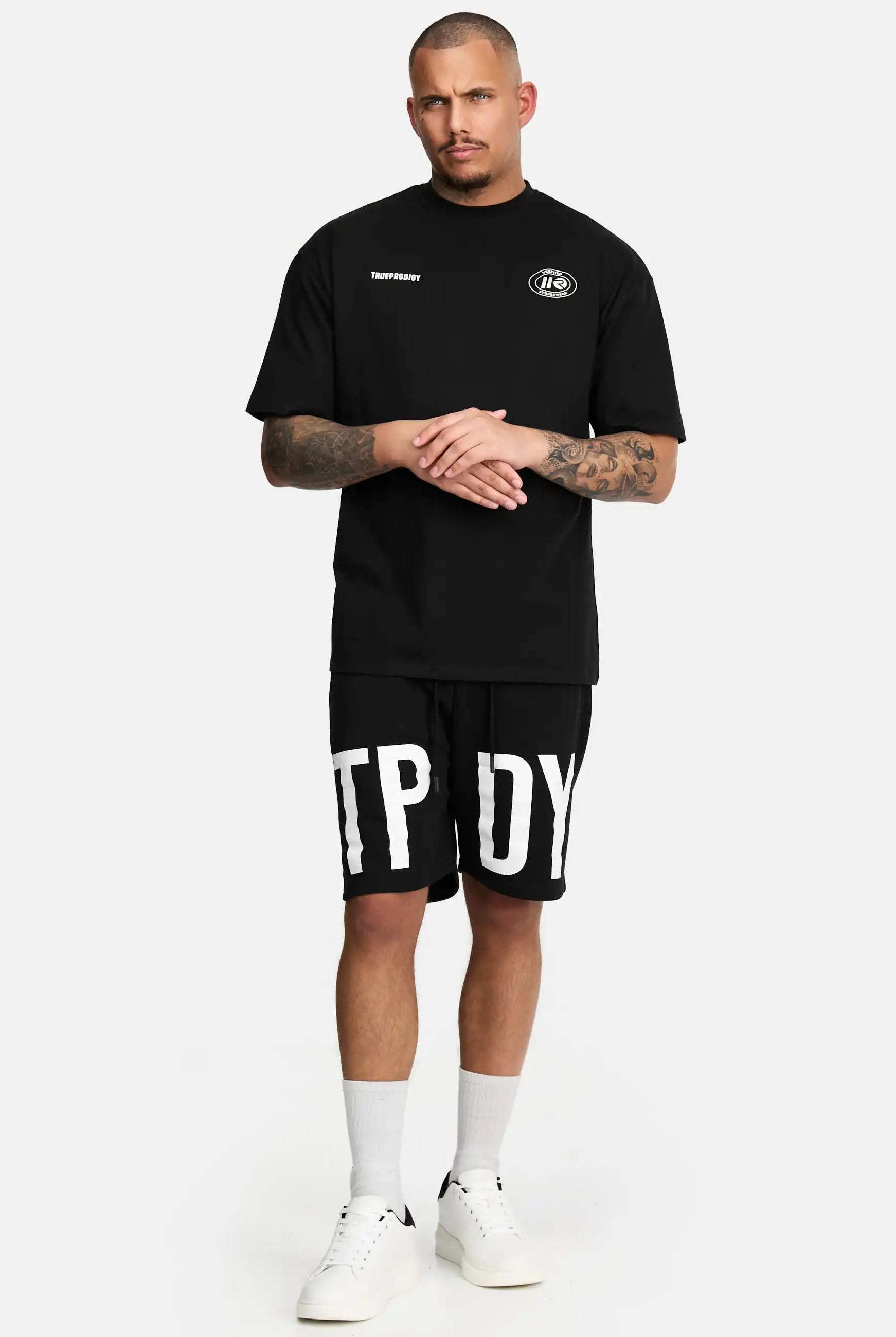 Ein Mann mit Tattoos und einem neutralen Gesichtsausdruck steht vor einem weißen Hintergrund und trägt das trueprodigy Channing Oversized T-Shirt mit Druck, schwarze Shorts mit fetten weißen Buchstaben, weiße Socken und Turnschuhe.
