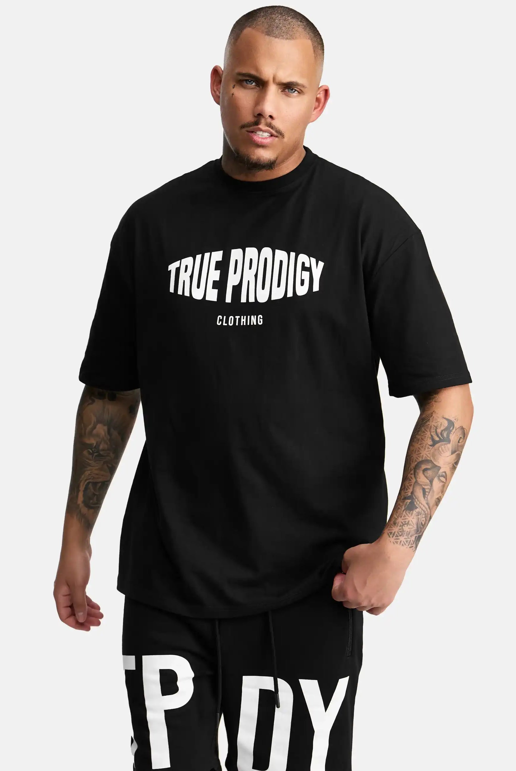 Ein tätowierter Mann trägt das Miguel Oversized T-Shirt mit Druck von trueprodigy, mit auffälligem weißem Druck, gepaart mit einer passenden schwarzen Hose mit großen weißen Buchstaben, vor einem schlichten hellen Hintergrund.