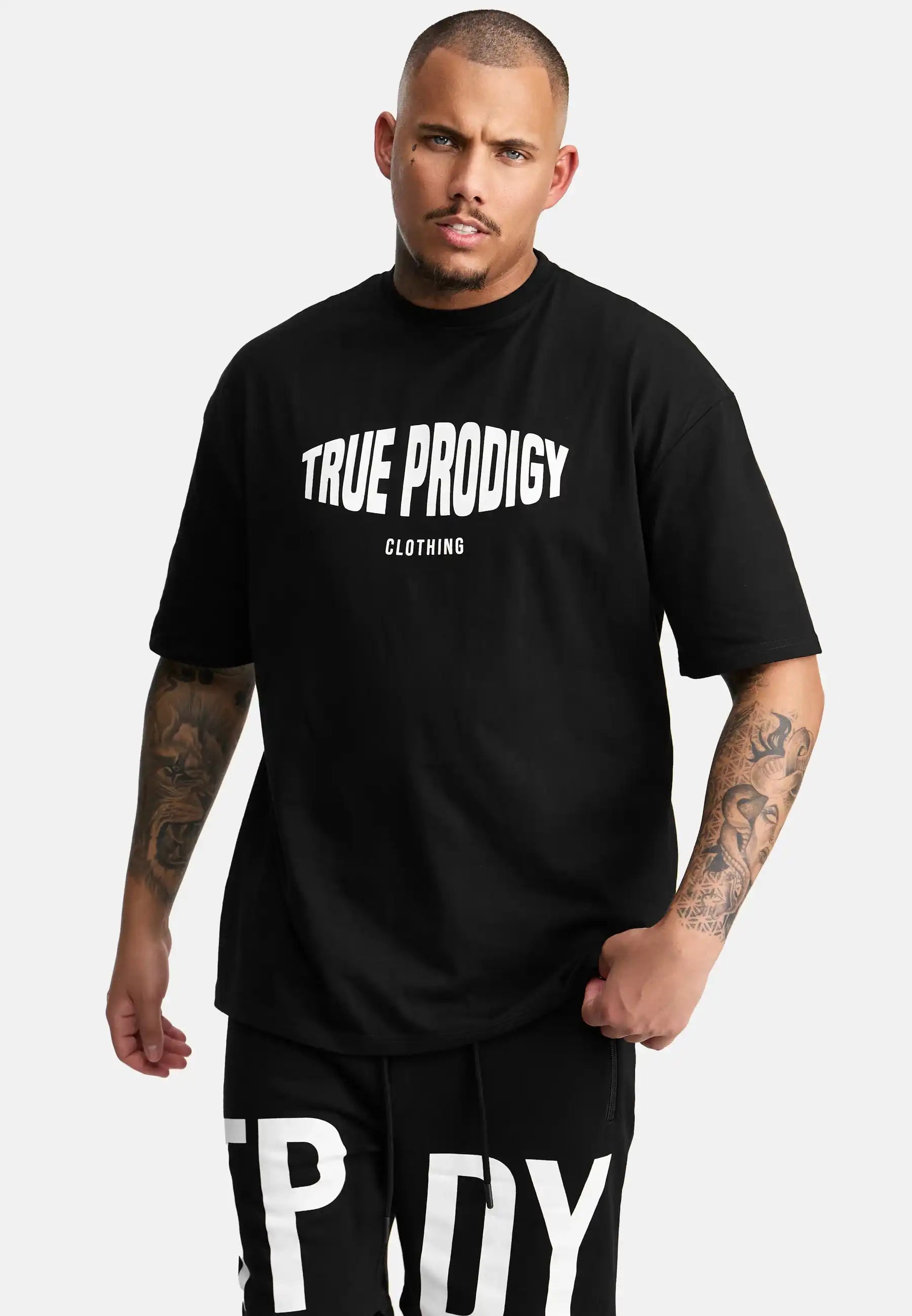 Ein tätowierter Mann trägt das Miguel Oversized T-Shirt mit Druck von trueprodigy, mit auffälligem weißem Druck, gepaart mit einer passenden schwarzen Hose mit großen weißen Buchstaben, vor einem schlichten hellen Hintergrund.