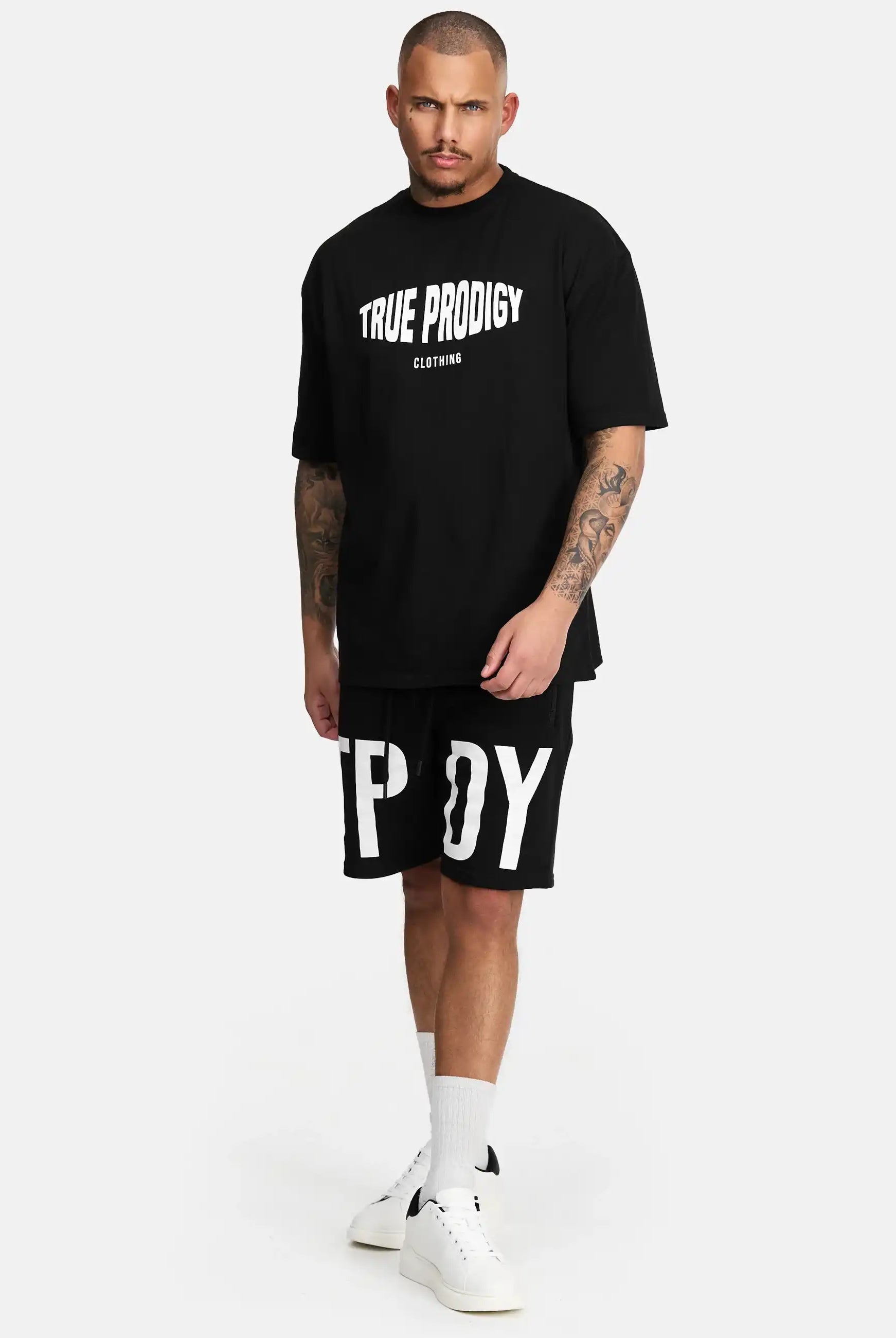 Ein Mann steht vor einem weißen Hintergrund und trägt das trueprodigy Miguel Oversized T-Shirt mit Druck, schwere schwarze Shorts mit großen weißen Buchstaben, weiße Socken und weiße Turnschuhe.