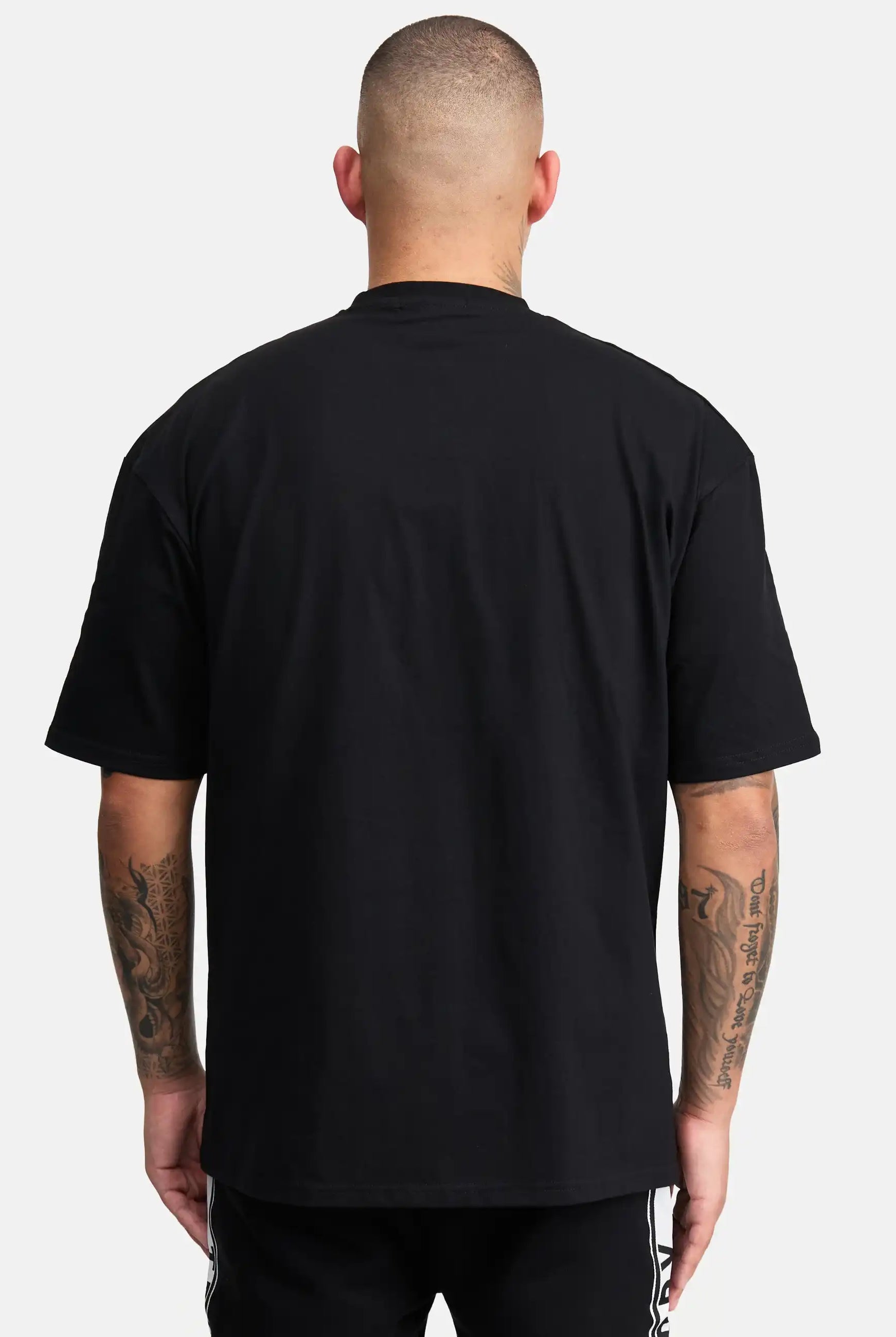 Eine Person mit kurzen Haaren, von hinten gesehen vor einem weißen Hintergrund, trägt ein schwarzes Cornelius Oversized T-Shirt mit Druck von trueprodigy und eine schwarze Hose. Ihre tätowierten Arme sind sichtbar.