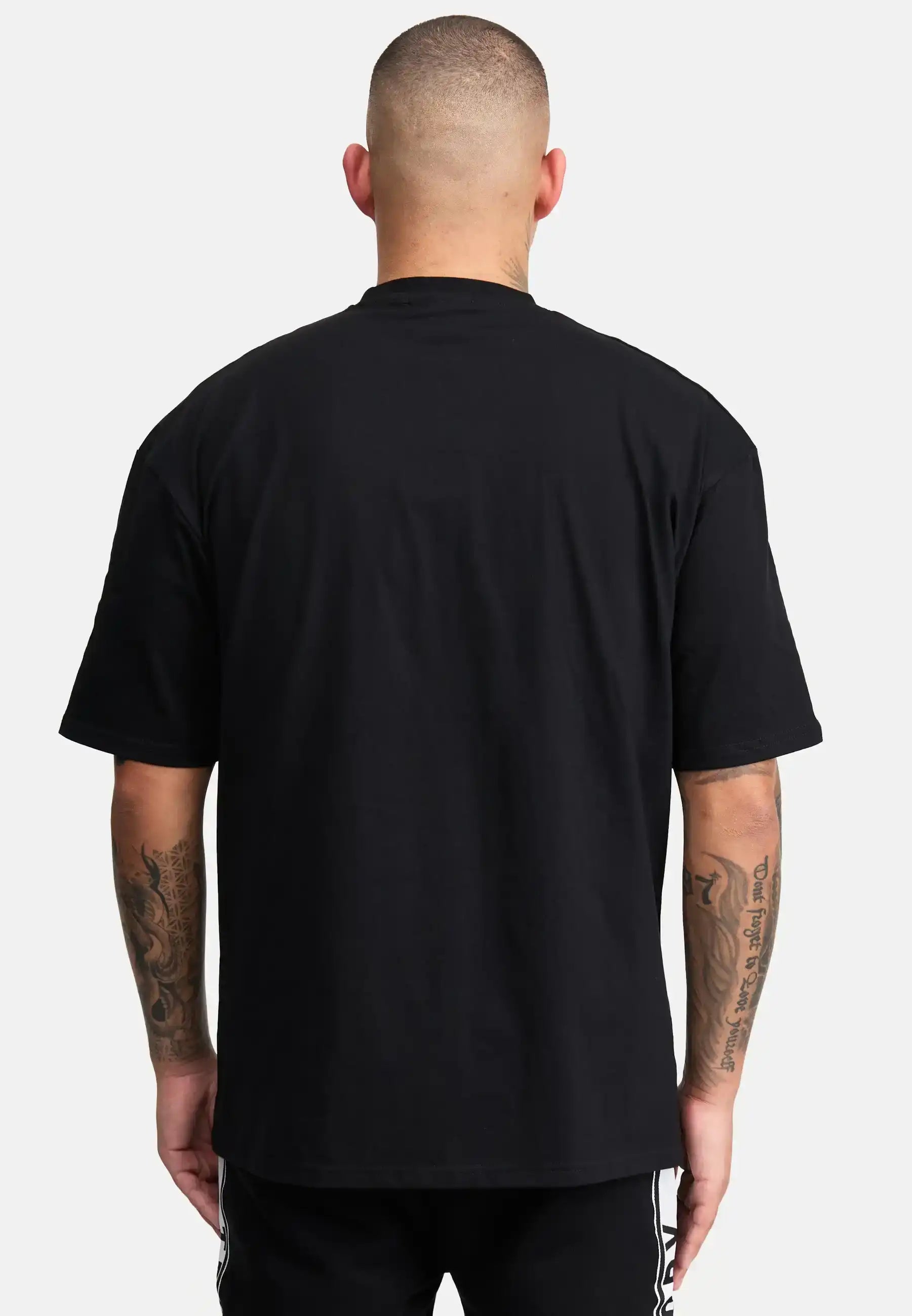 Eine Person mit kurzen Haaren, von hinten gesehen vor einem weißen Hintergrund, trägt ein schwarzes Cornelius Oversized T-Shirt mit Druck von trueprodigy und eine schwarze Hose. Ihre tätowierten Arme sind sichtbar.