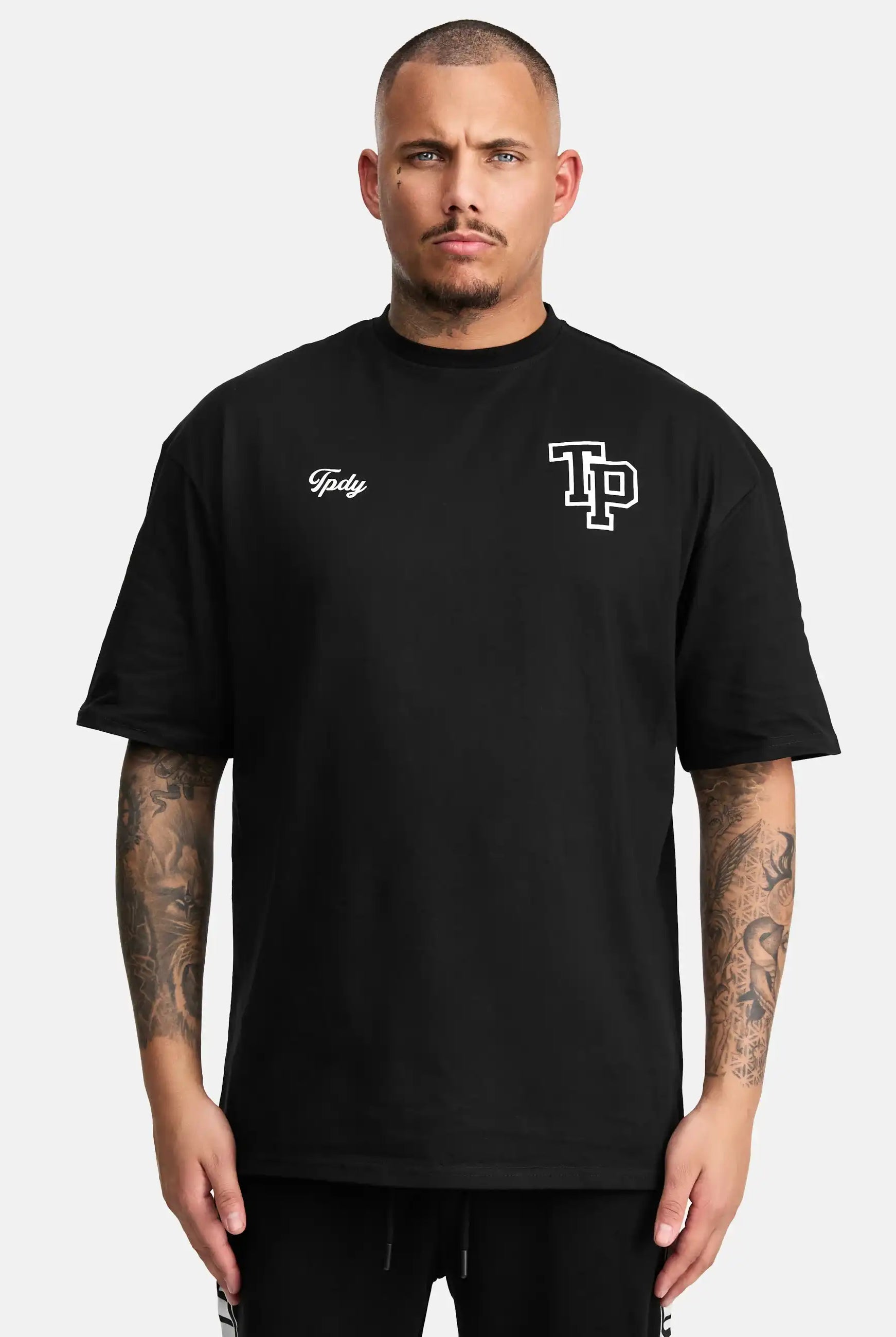 Eine Person mit kurzen Haaren und Tattoos auf beiden Armen trägt das trueprodigy Cornelius Oversized T-Shirt mit Druck, ein lockeres Unisex-T-Shirt aus schwerem Jersey, auf dem TP und Toby in weißer Farbe auf der Vorderseite abgebildet sind, vor einem schlichten weißen Hintergrund.