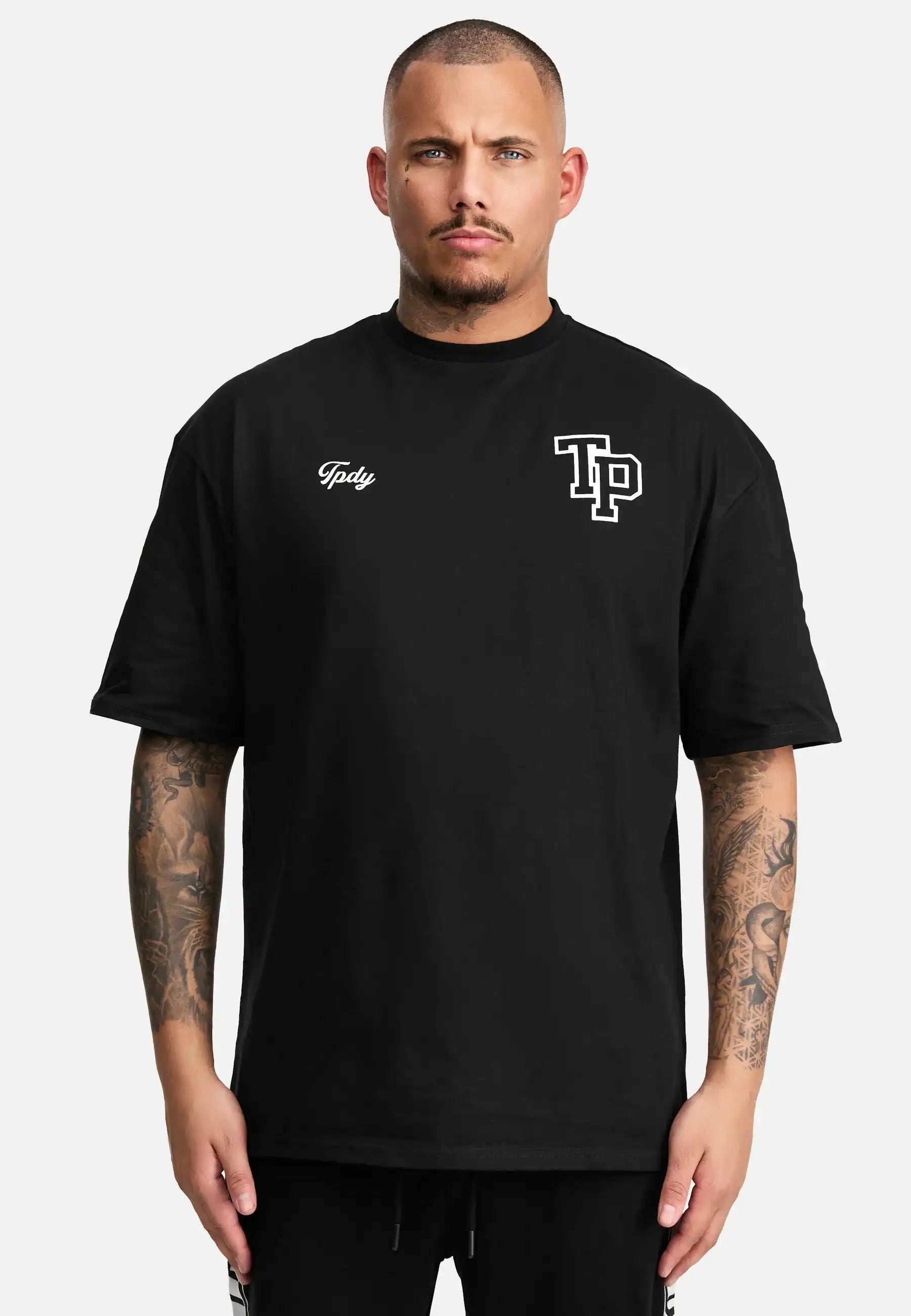 Eine Person mit kurzen Haaren und Tattoos auf beiden Armen trägt das trueprodigy Cornelius Oversized T-Shirt mit Druck, ein lockeres Unisex-T-Shirt aus schwerem Jersey, auf dem TP und Toby in weißer Farbe auf der Vorderseite abgebildet sind, vor einem schlichten weißen Hintergrund.
