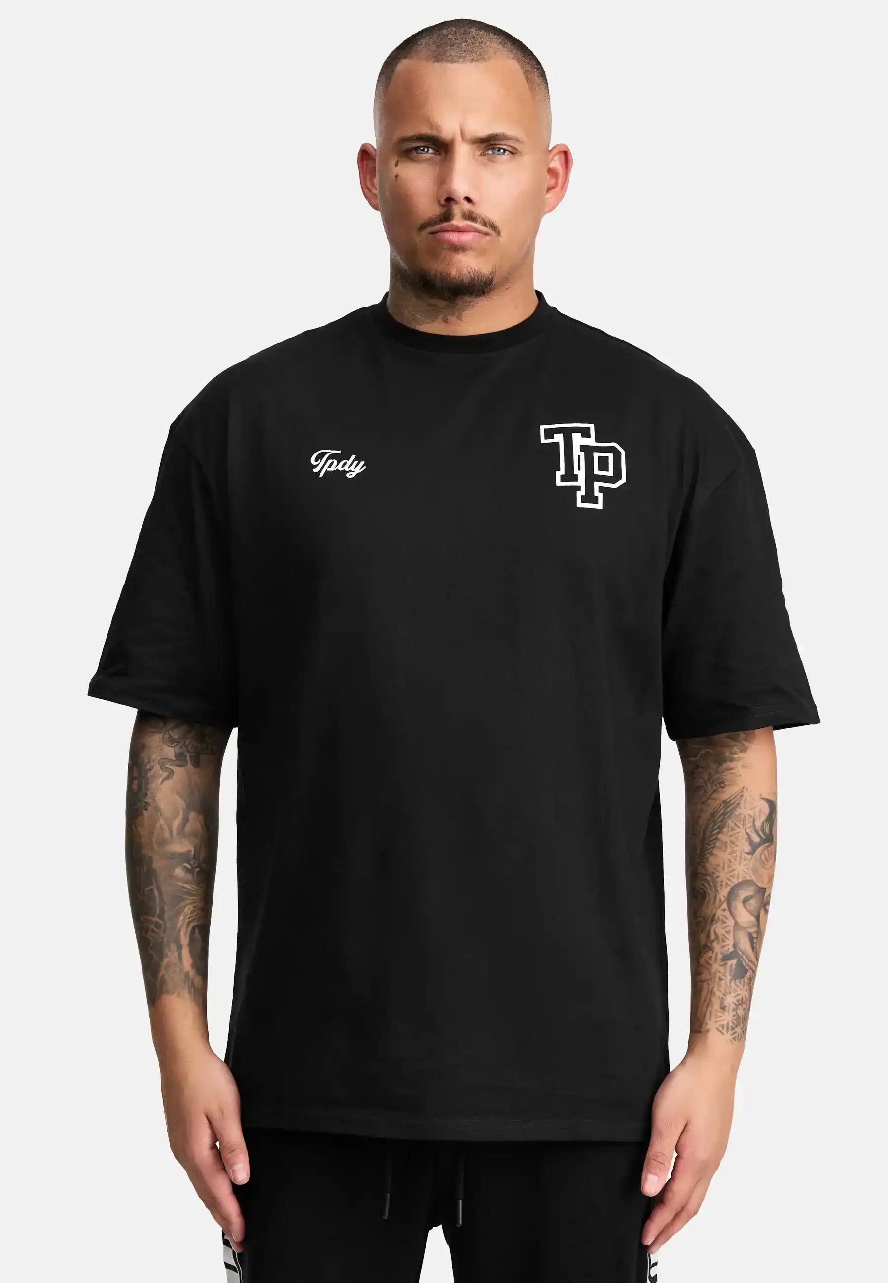 Eine Person mit kurzen Haaren und Tattoos auf beiden Armen trägt das trueprodigy Cornelius Oversized T-Shirt mit Druck, ein lockeres Unisex-T-Shirt aus schwerem Jersey, auf dem TP und Toby in weißer Farbe auf der Vorderseite abgebildet sind, vor einem schlichten weißen Hintergrund.