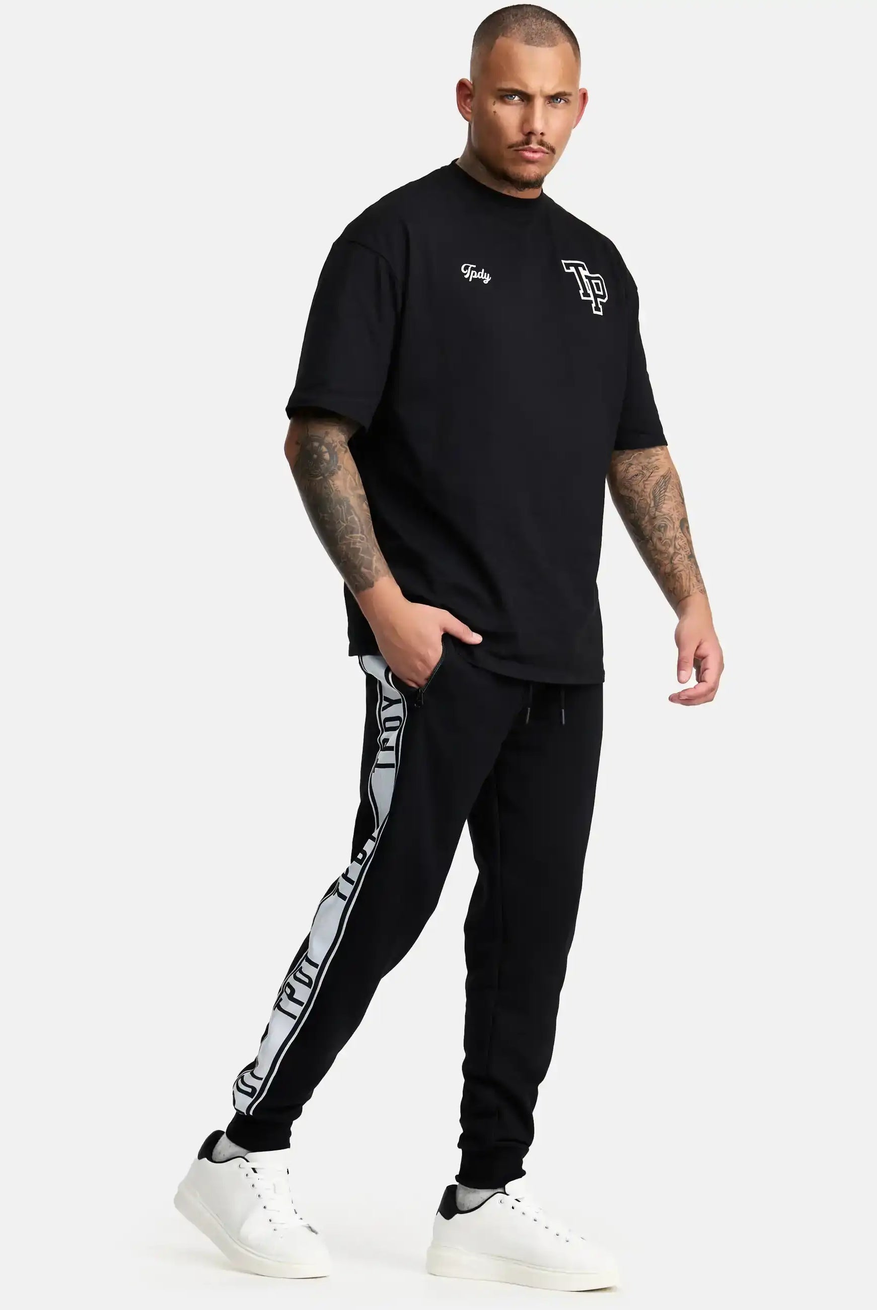 Ein Mann mit Tattoos und Kurzhaarschnitt steht vor einem schlichten Hintergrund und trägt das trueprodigy Cornelius Oversized T-Shirt mit Druck, eine schwarze Trainingshose mit weißen Logostreifen und weiße Turnschuhe.