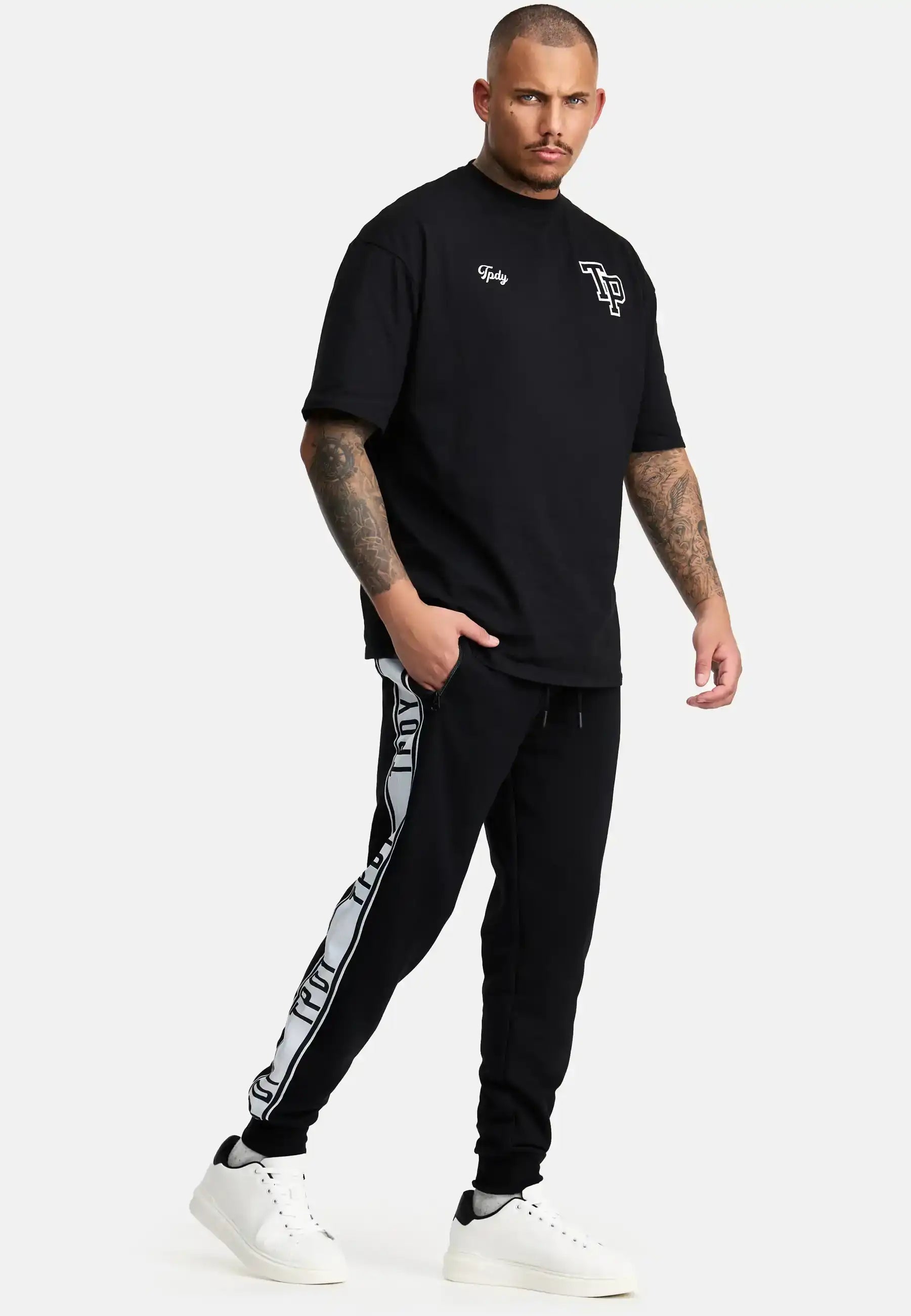 Ein Mann mit Tattoos und Kurzhaarschnitt steht vor einem schlichten Hintergrund und trägt das trueprodigy Cornelius Oversized T-Shirt mit Druck, eine schwarze Trainingshose mit weißen Logostreifen und weiße Turnschuhe.