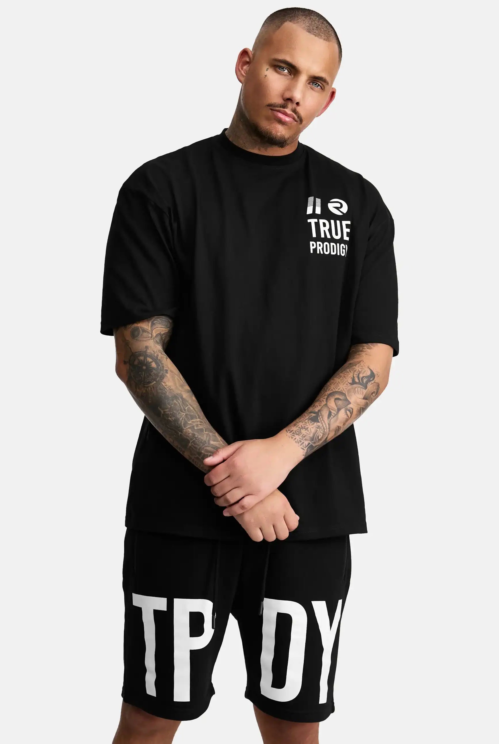 Ein tätowierter Mann mit kurzen Haaren und gestutztem Bart steht vor einem schlichten Hintergrund. Er trägt ein schwarzes trueprodigy Peter Oversized T-Shirt mit Druck und schwarze Shorts mit großen weißen TPDY Buchstaben.