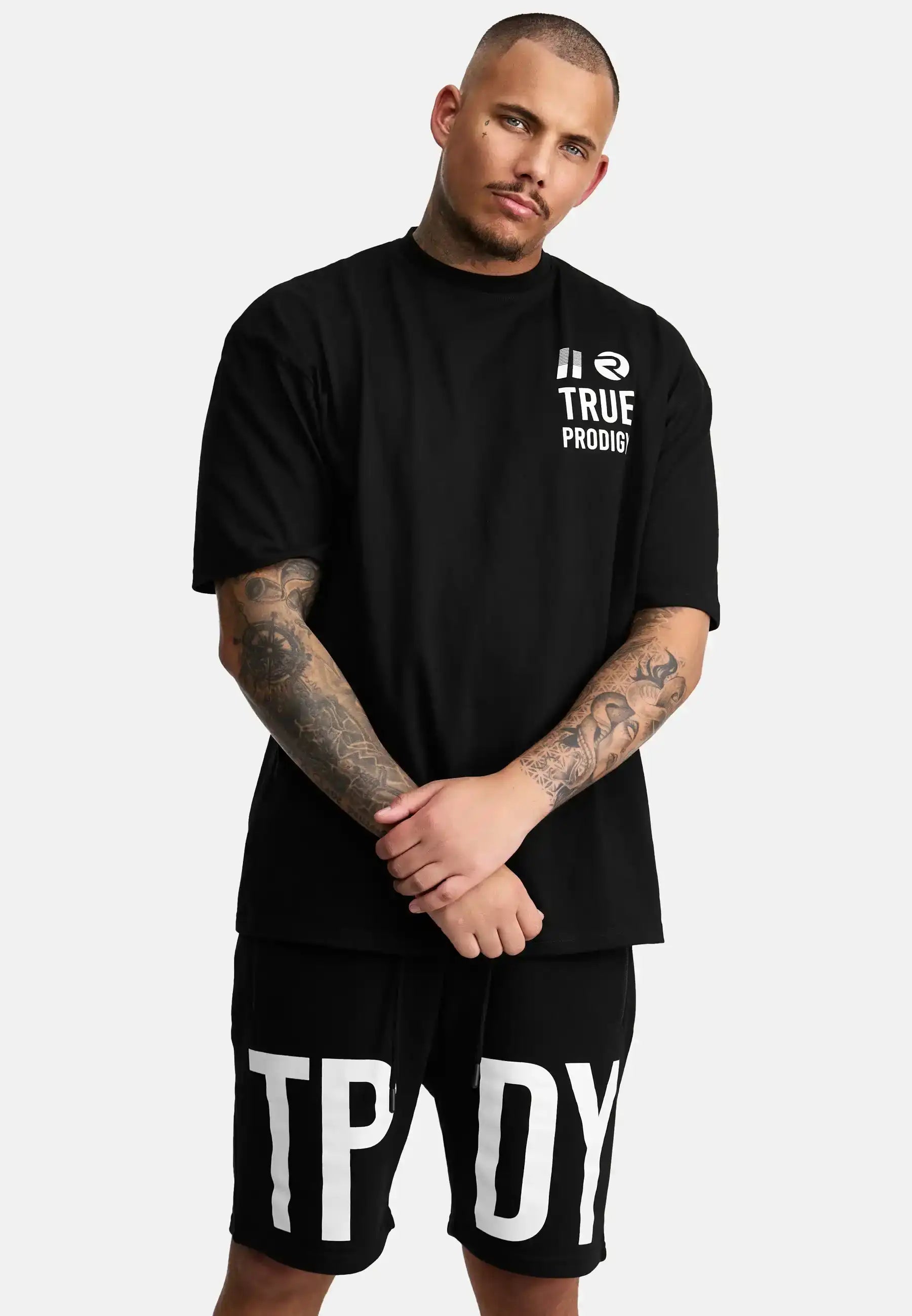 Ein tätowierter Mann mit kurzen Haaren und gestutztem Bart steht vor einem schlichten Hintergrund. Er trägt ein schwarzes trueprodigy Peter Oversized T-Shirt mit Druck und schwarze Shorts mit großen weißen TPDY Buchstaben.