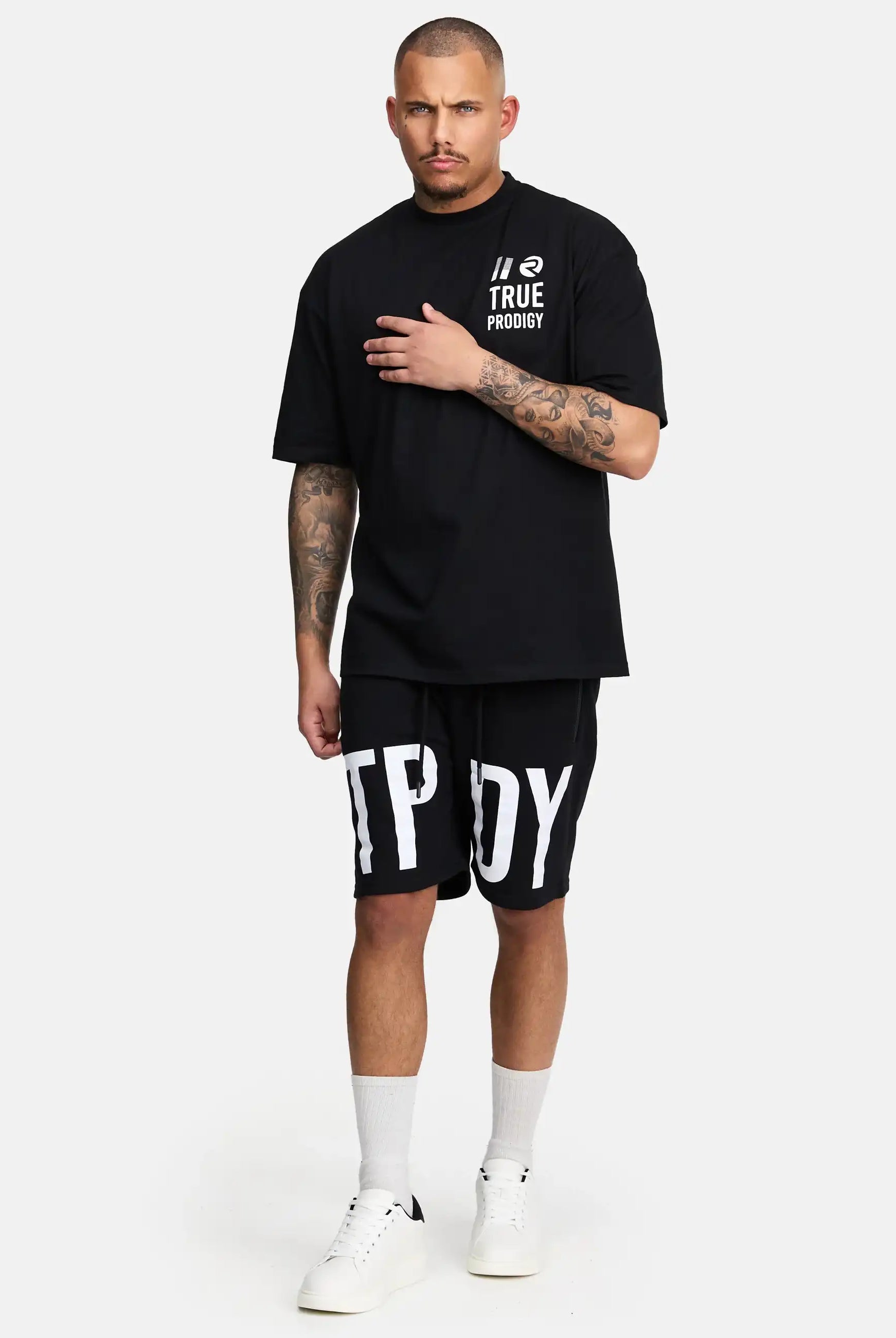 Ein tätowierter Mann steht vor einem schlichten Hintergrund und trägt ein schwarzes trueprodigy Peter Oversized T-Shirt mit Druck und passende schwarze Shorts mit großen weißen Buchstaben, dazu weiße Socken und Turnschuhe. Eine Hand ruht auf seiner Brust.