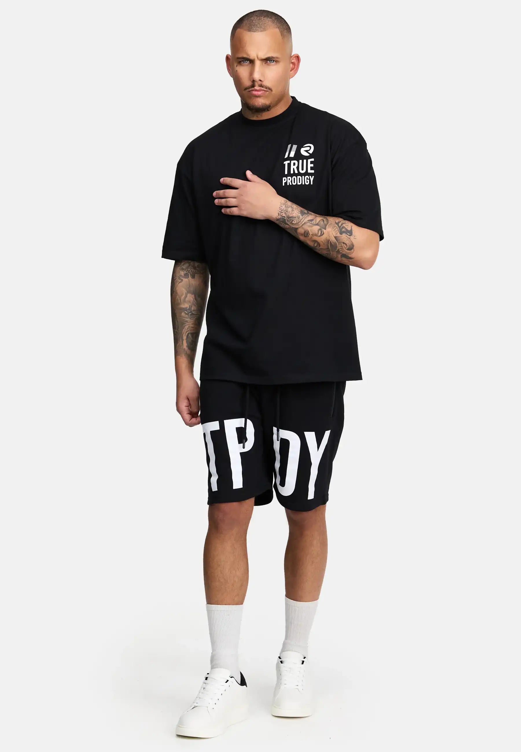 Ein tätowierter Mann steht vor einem schlichten Hintergrund und trägt ein schwarzes trueprodigy Peter Oversized T-Shirt mit Druck und passende schwarze Shorts mit großen weißen Buchstaben, dazu weiße Socken und Turnschuhe. Eine Hand ruht auf seiner Brust.