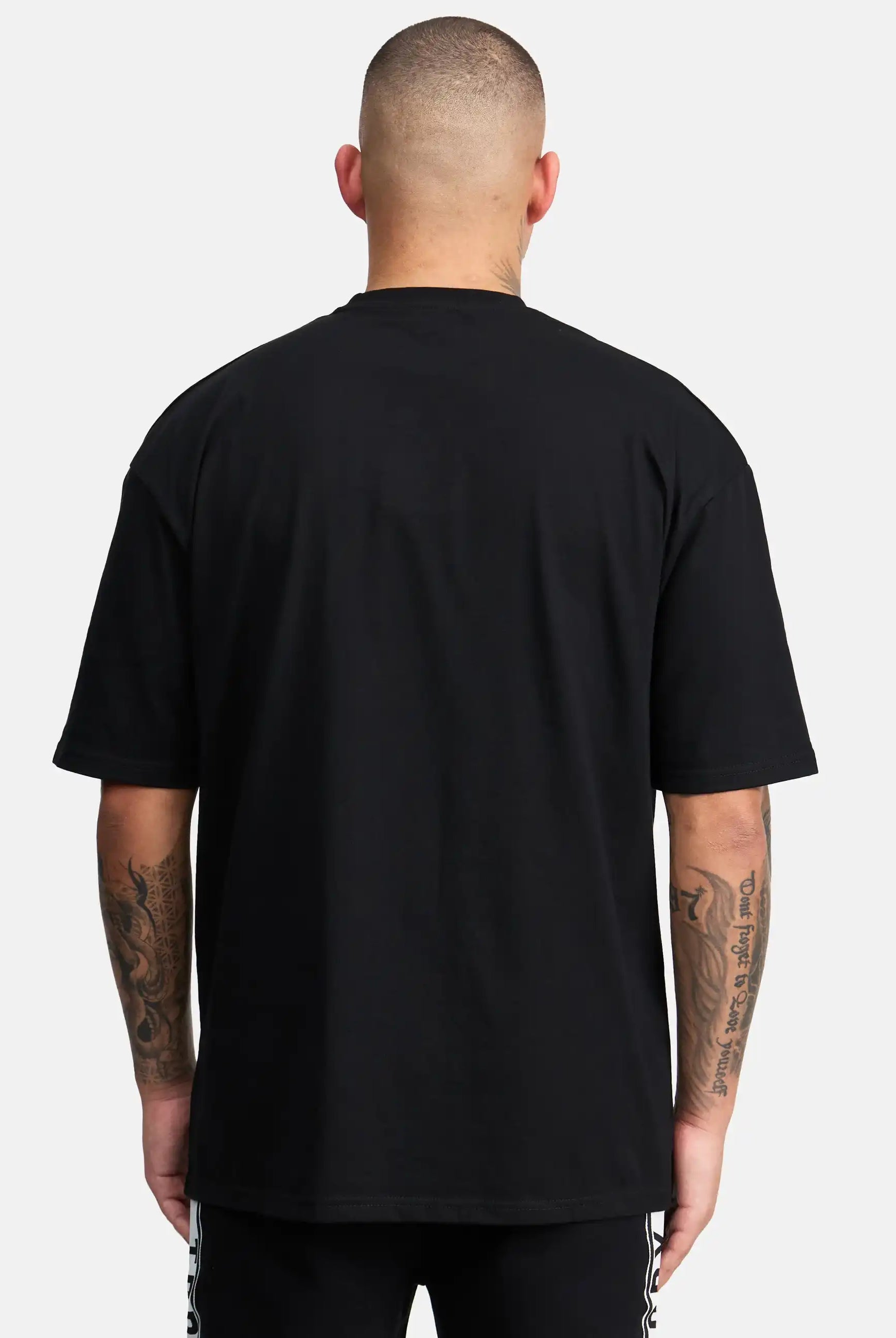 Eine Person mit kurzgeschnittenen Haaren und tätowierten Armen ist von hinten zu sehen. Sie trägt das trueprodigy Shaquell Oversized T-Shirt mit Druck und eine schwarze Hose, wobei sich die lockere Passform von einem schlichten weißen Hintergrund abhebt.