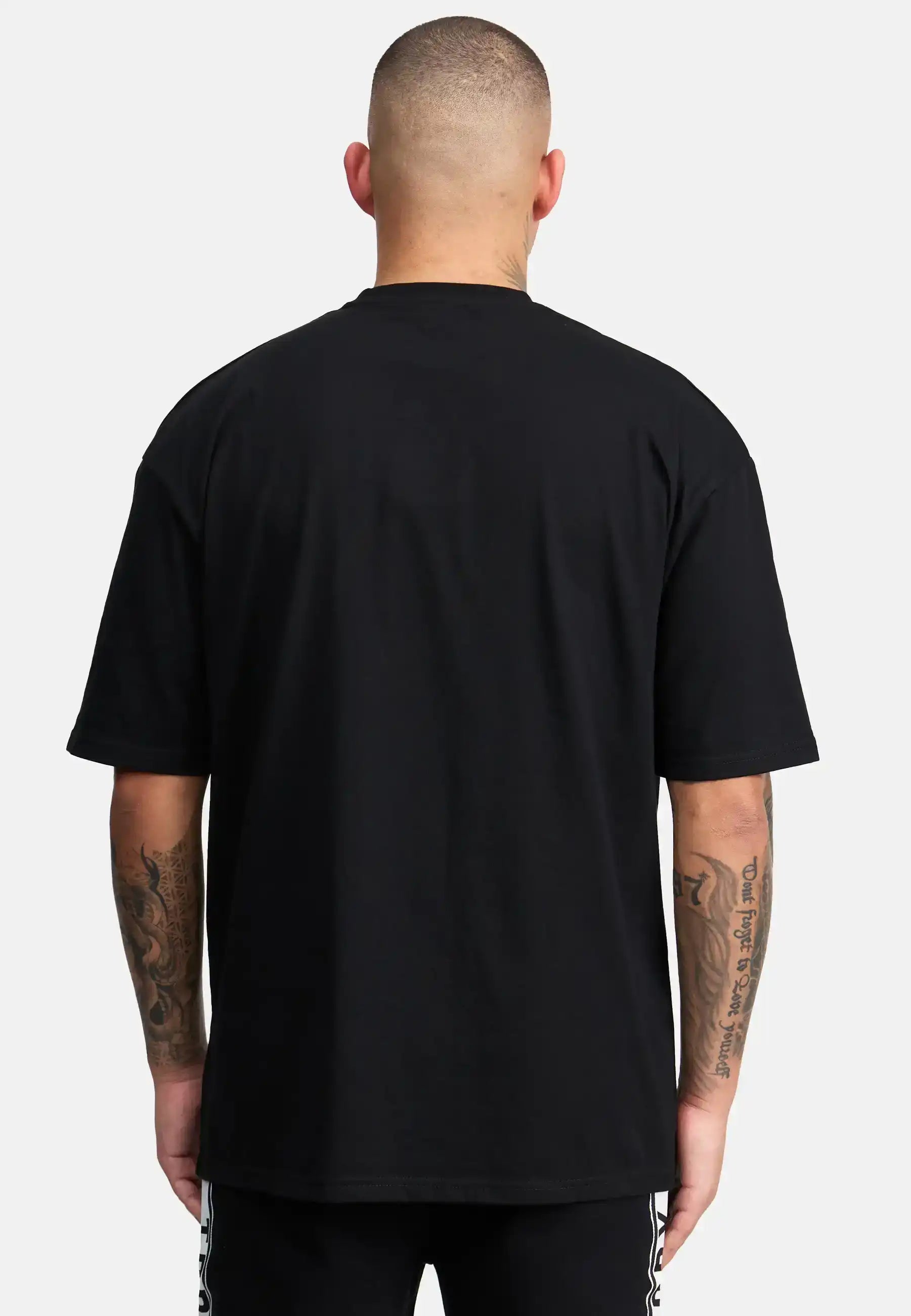 Eine Person mit kurzgeschnittenen Haaren und tätowierten Armen ist von hinten zu sehen. Sie trägt das trueprodigy Shaquell Oversized T-Shirt mit Druck und eine schwarze Hose, wobei sich die lockere Passform von einem schlichten weißen Hintergrund abhebt.