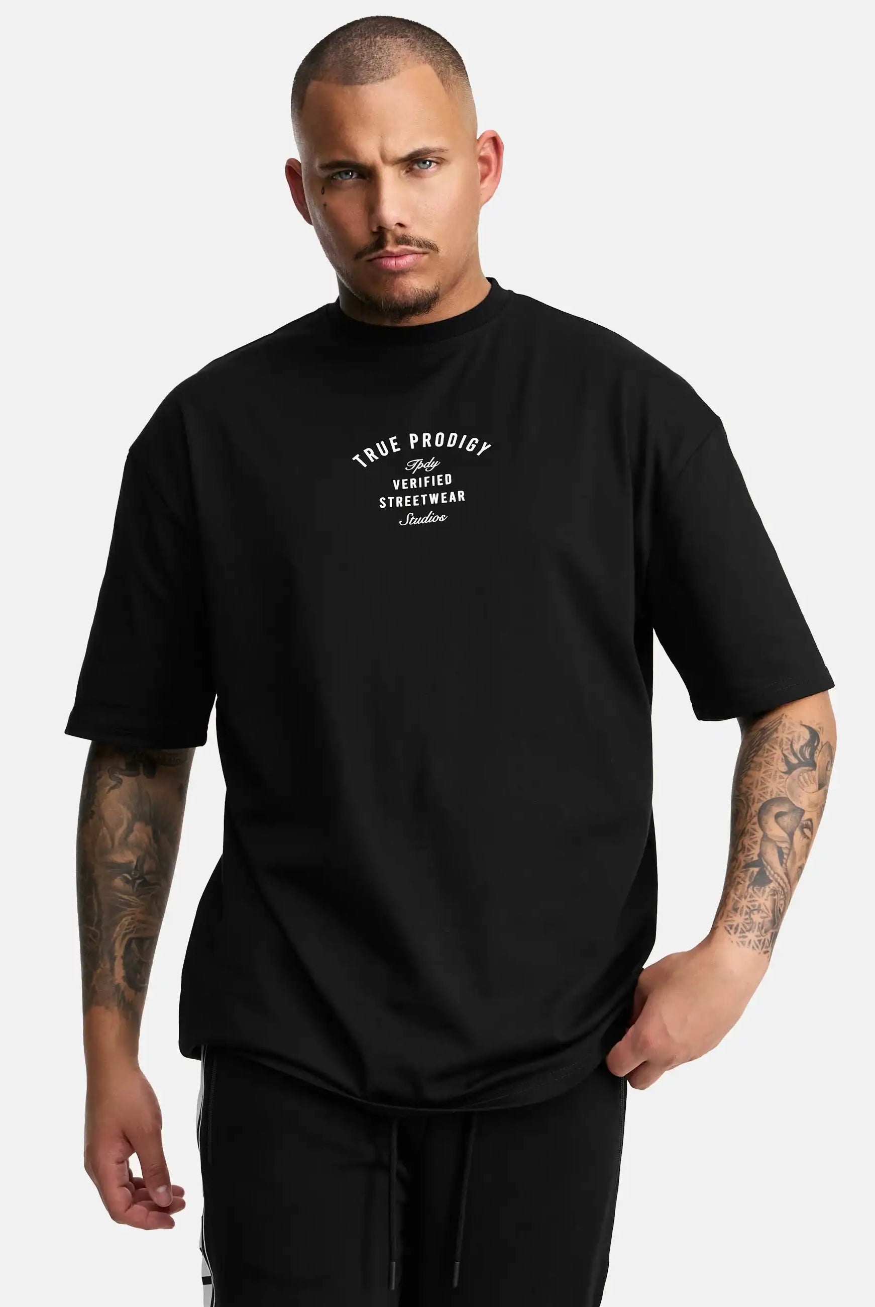 Eine tätowierte Person mit kurzgeschorenen Haaren trägt das trueprodigy Shaquell Oversized T-Shirt mit Druck in schwarz mit weißem "True Prodigy Verified Streetwear London"-Text, vor einem schlichten weißen Hintergrund.