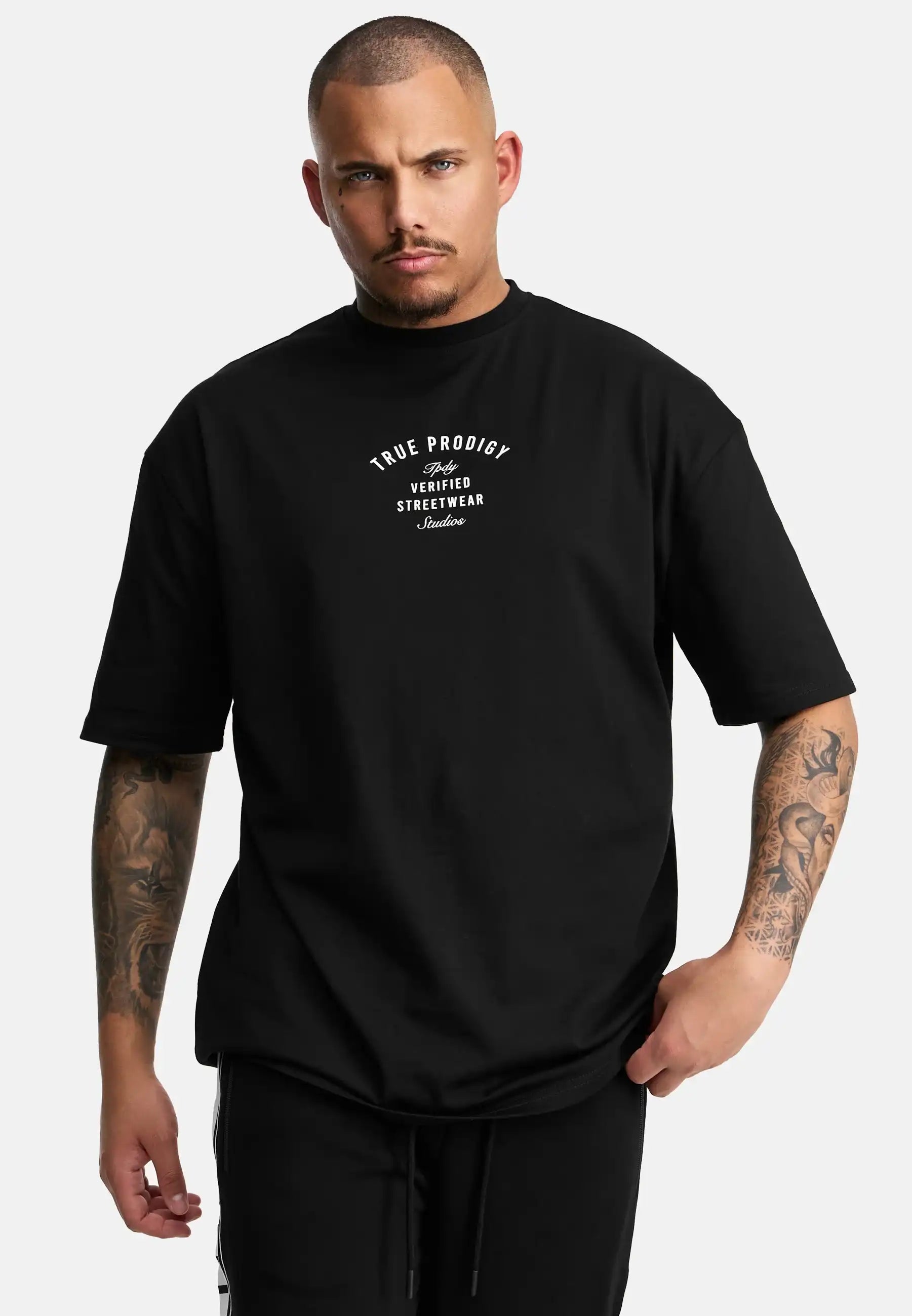 Eine tätowierte Person mit kurzgeschorenen Haaren trägt das trueprodigy Shaquell Oversized T-Shirt mit Druck in schwarz mit weißem "True Prodigy Verified Streetwear London"-Text, vor einem schlichten weißen Hintergrund.