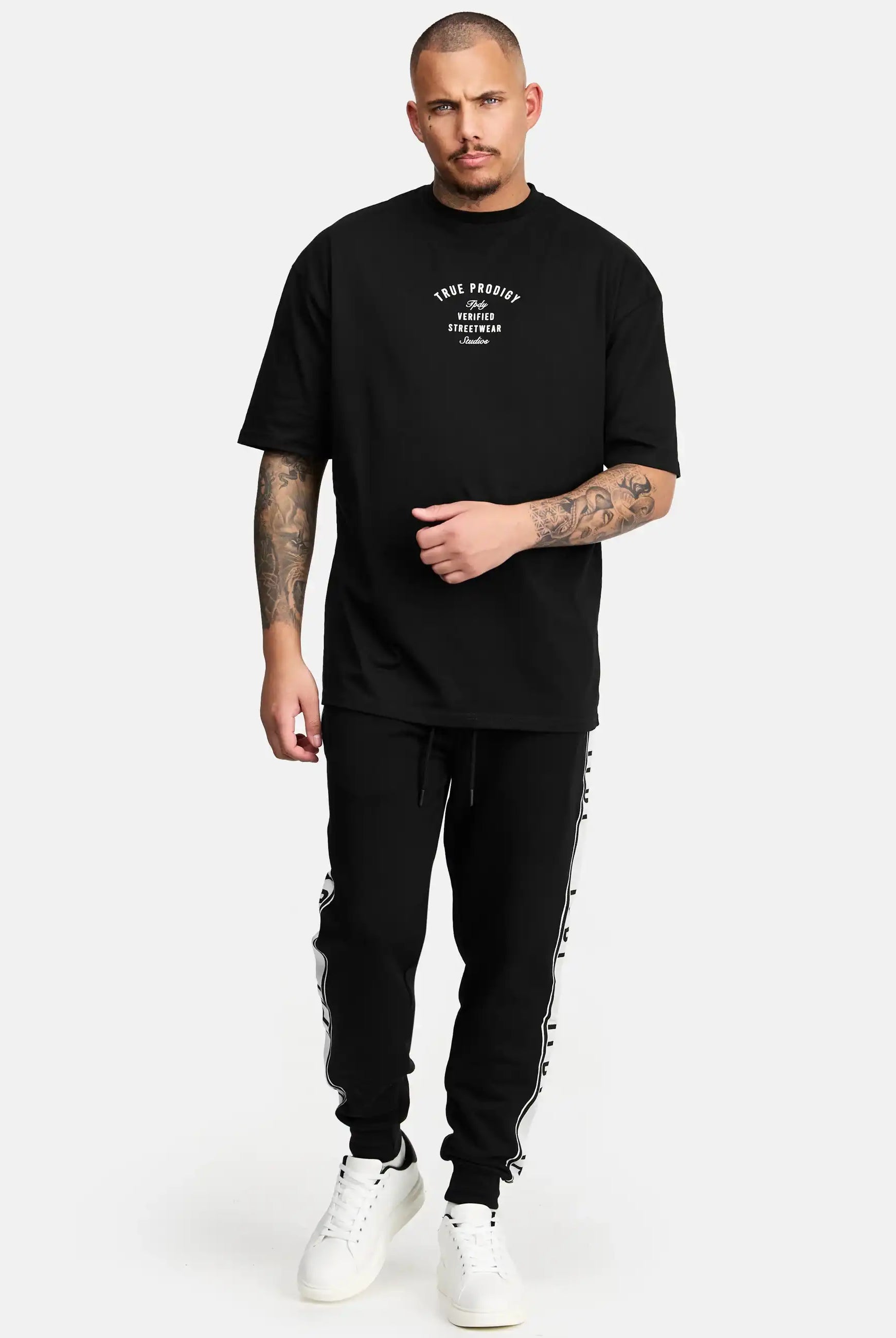 Ein Mann mit kurzen Haaren und Tattoos steht vor einem schlichten Hintergrund und trägt ein schwarzes trueprodigy Shaquell Oversized T-Shirt mit Druck, eine schwarze Trainingshose mit Seitenstreifen und weiße Turnschuhe.