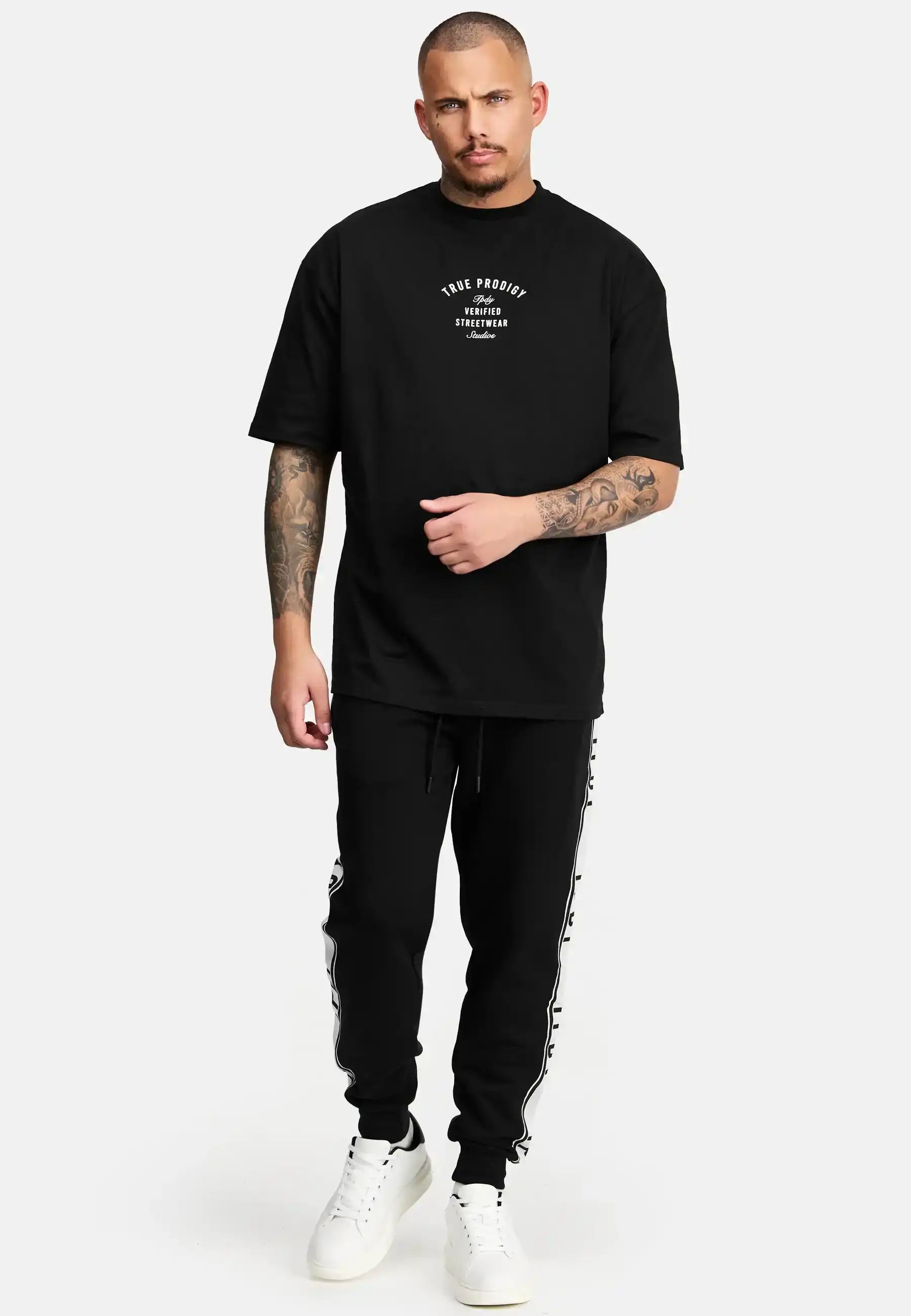 Ein Mann mit kurzen Haaren und Tattoos steht vor einem schlichten Hintergrund und trägt ein schwarzes trueprodigy Shaquell Oversized T-Shirt mit Druck, eine schwarze Trainingshose mit Seitenstreifen und weiße Turnschuhe.
