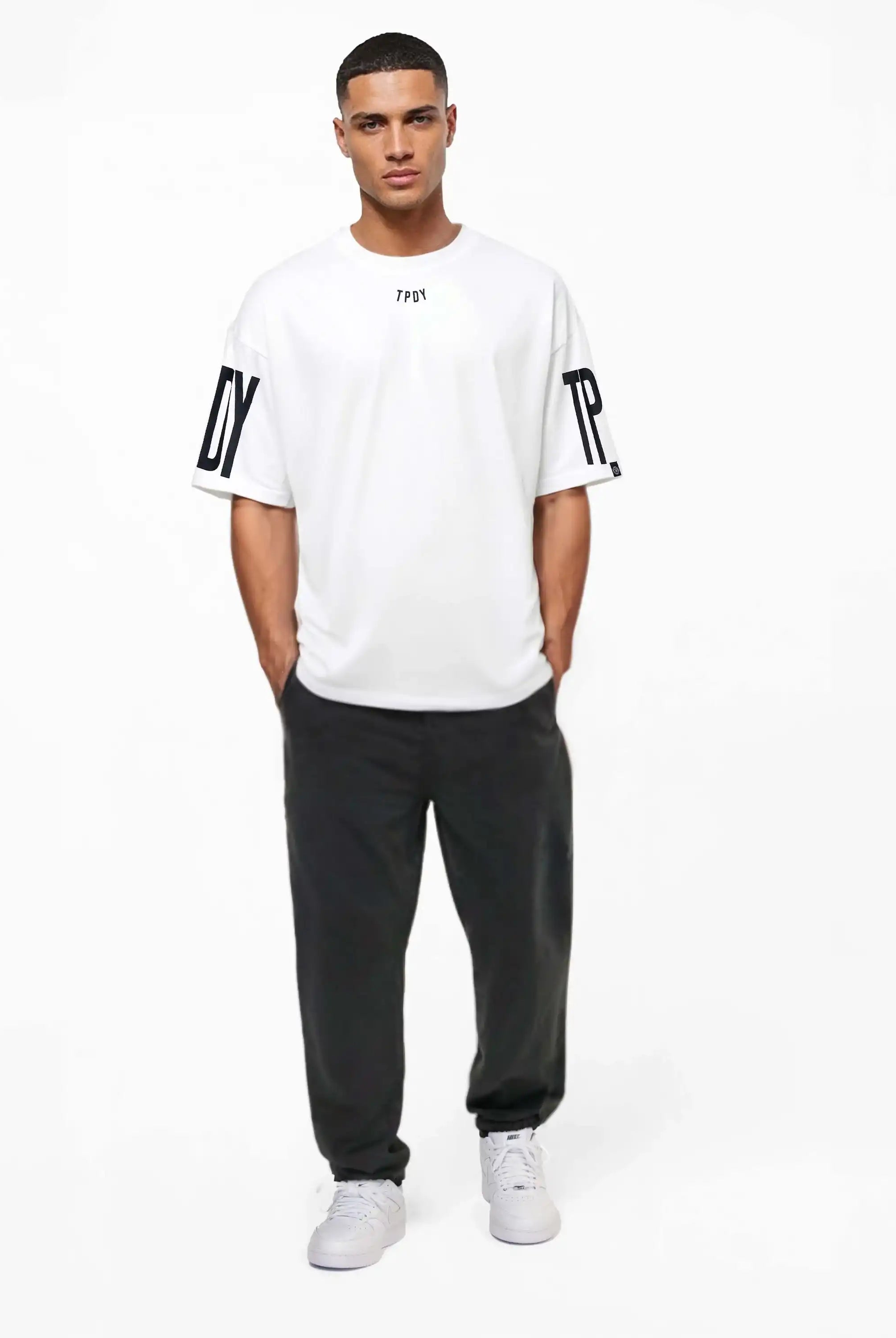 Ein Mann blickt nach vorne und trägt das trueprodigy Viktor Oversize-Herren-T-Shirt mit Front- und Ärmelprints, gepaart mit einer schwarzen Hose und weißen Turnschuhen, vor einem schlichten weißen Hintergrund.