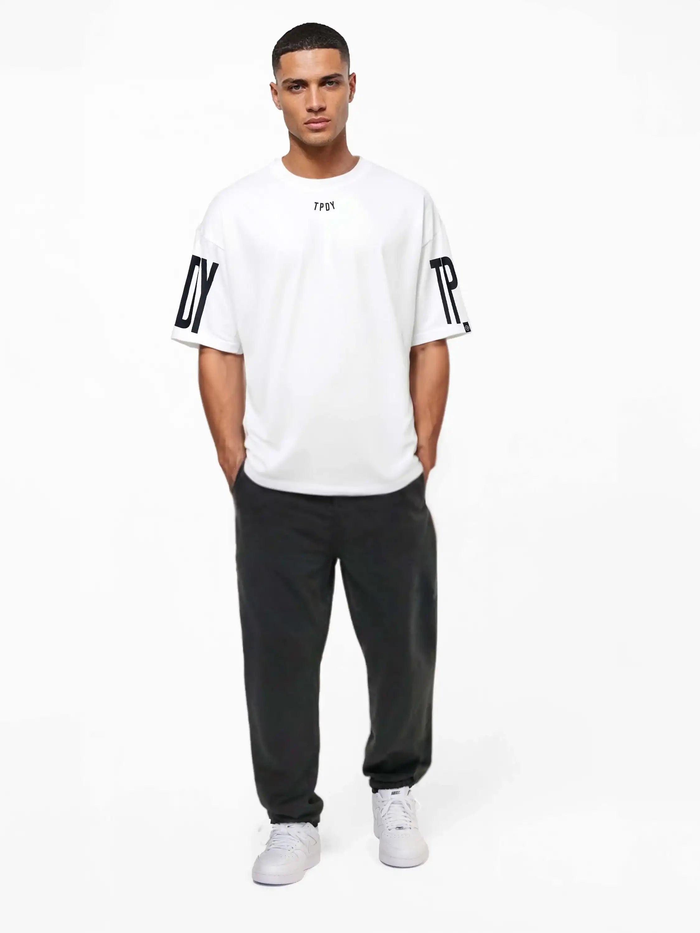 Ein Mann blickt nach vorne und trägt das trueprodigy Viktor Oversize-Herren-T-Shirt mit Front- und Ärmelprints, gepaart mit einer schwarzen Hose und weißen Turnschuhen, vor einem schlichten weißen Hintergrund.