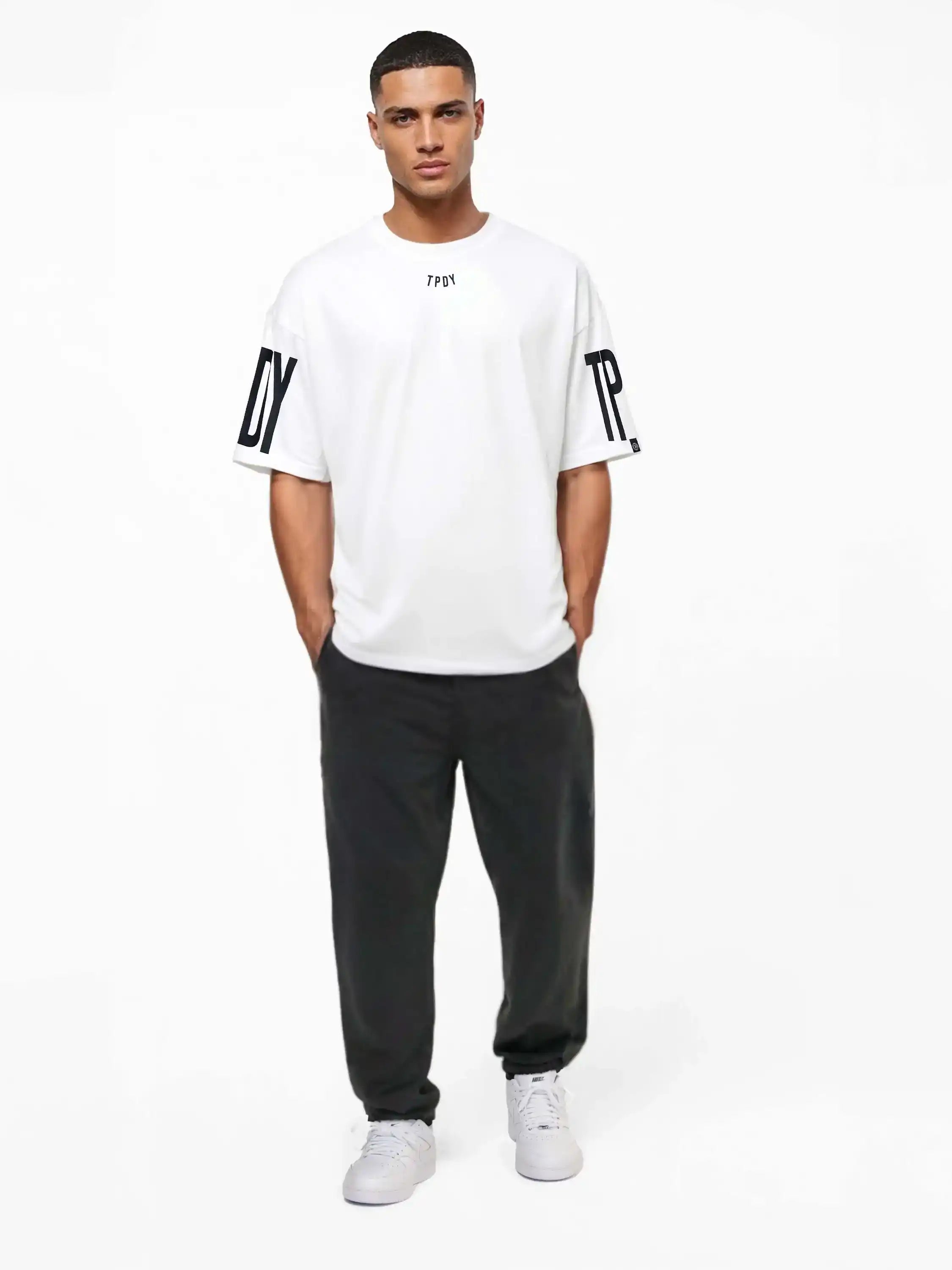 Ein Mann blickt nach vorne und trägt das trueprodigy Viktor Oversize-Herren-T-Shirt mit Front- und Ärmelprints, gepaart mit einer schwarzen Hose und weißen Turnschuhen, vor einem schlichten weißen Hintergrund.