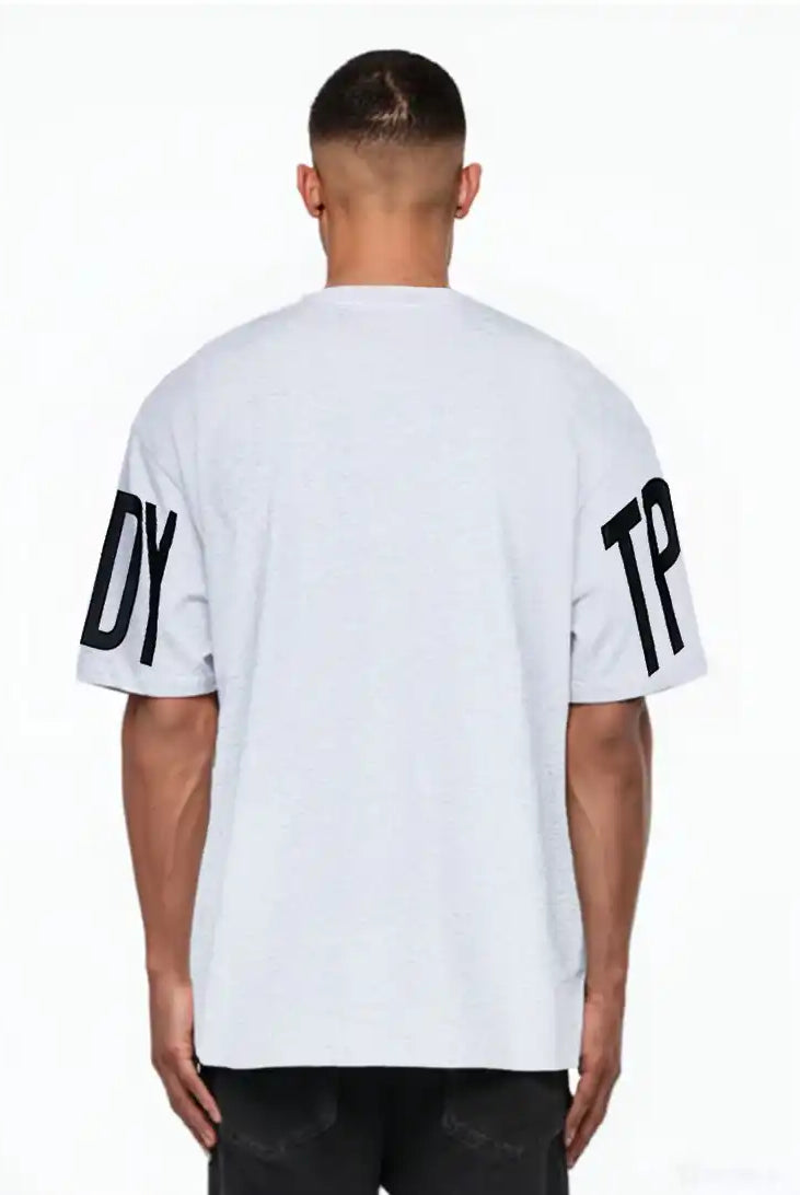 Eine Person schaut von der Kamera weg und trägt das trueprodigy Viktor Oversize-Herren-T-Shirt mit Front- und Ärmelprints in Hellgrau mit auffälligen schwarzen DY- und TP-Ärmelprints, gepaart mit einer dunklen Hose.