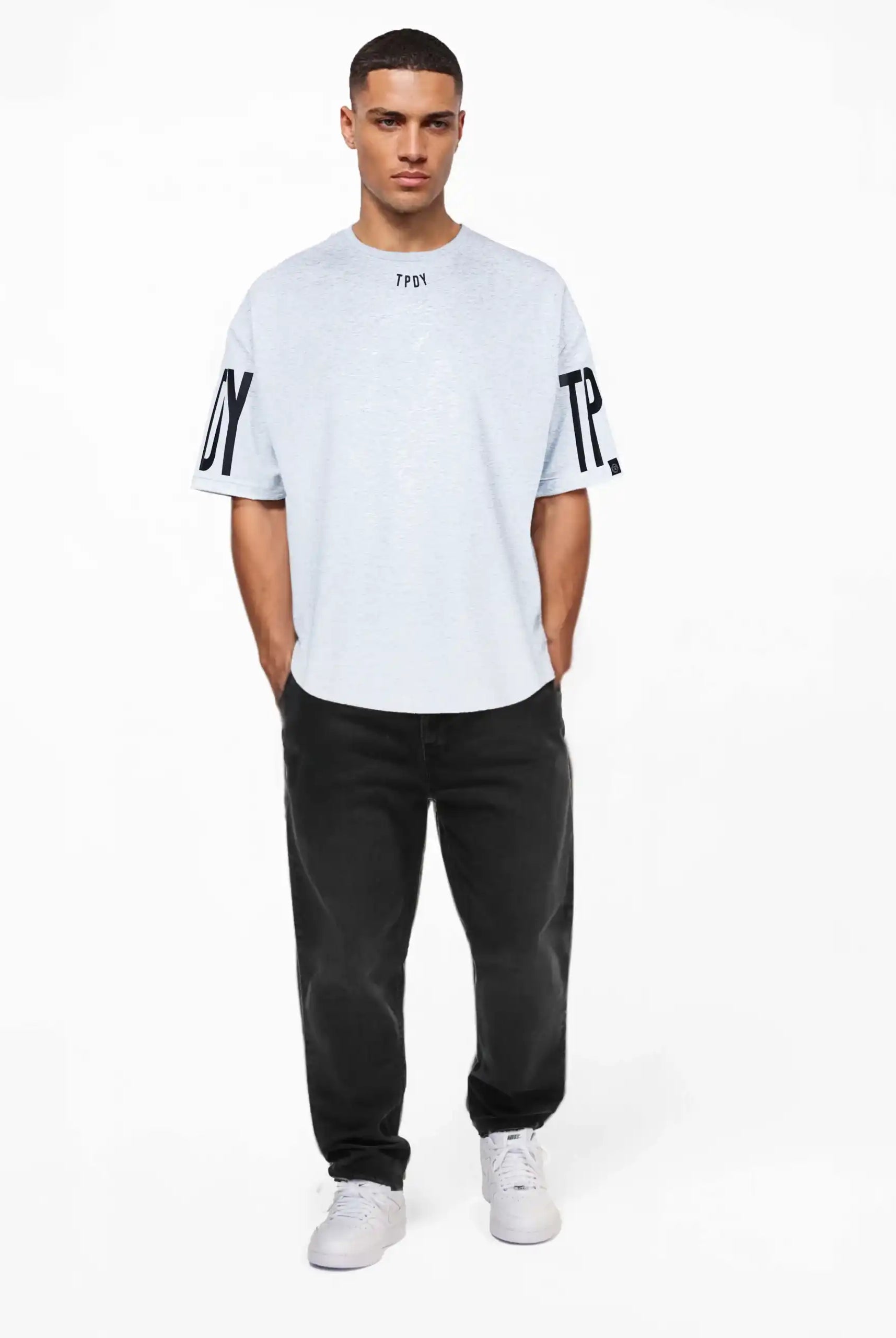 Ein Mann trägt das Viktor Oversize-Herren-T-Shirt mit Front- und Ärmelprints von trueprodigy mit TPU-Text und schwarzen Ärmelprints, gepaart mit schwarzen Jeans und weißen Turnschuhen, Hände in den Taschen, vor einem schlichten weißen Hintergrund.