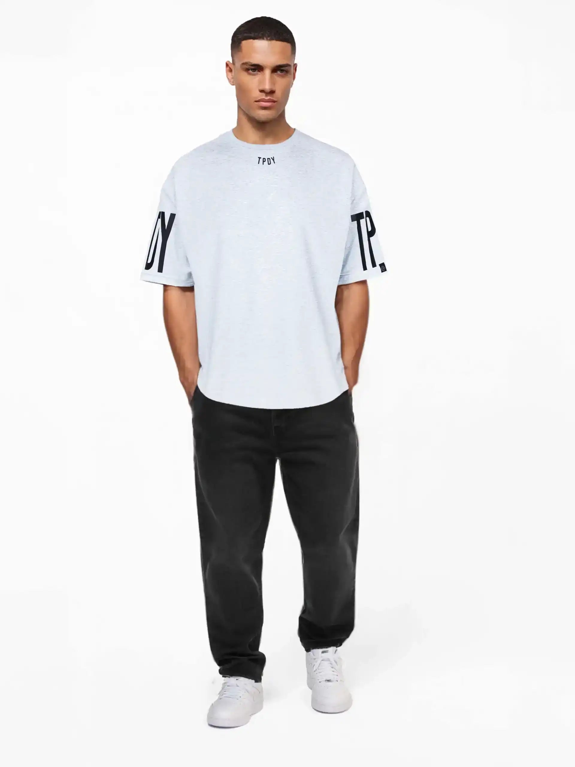 Ein Mann trägt das Viktor Oversize-Herren-T-Shirt mit Front- und Ärmelprints von trueprodigy mit TPU-Text und schwarzen Ärmelprints, gepaart mit schwarzen Jeans und weißen Turnschuhen, Hände in den Taschen, vor einem schlichten weißen Hintergrund.