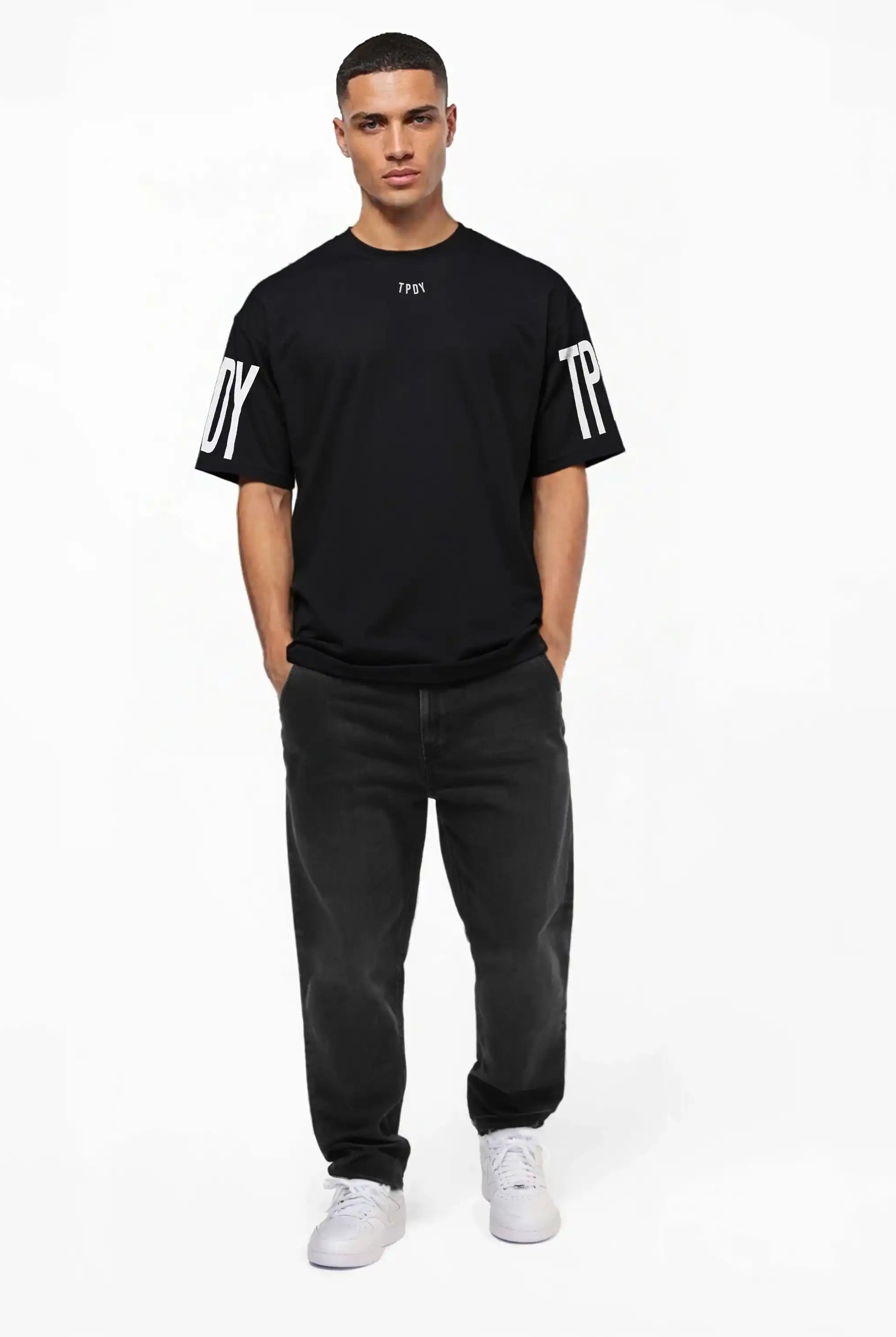 Ein Mann blickt mit den Händen in den Taschen nach vorne und trägt das trueprodigy Viktor Oversize-Herren-T-Shirt mit Front- und Ärmelprints, gepaart mit dunklen Jeans und weißen Turnschuhen vor einem schlichten weißen Hintergrund.