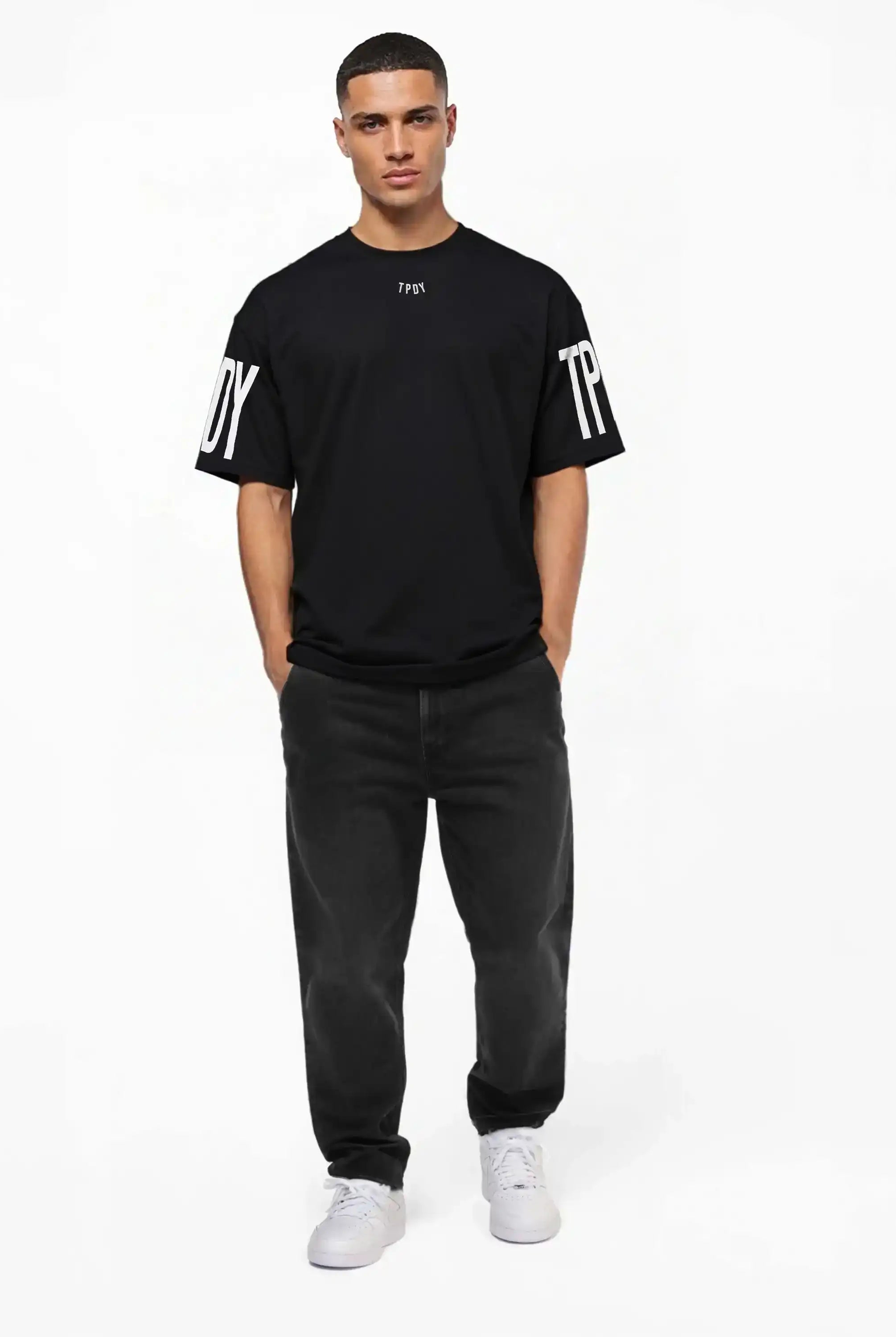 Ein Mann blickt mit den Händen in den Taschen nach vorne und trägt das trueprodigy Viktor Oversize-Herren-T-Shirt mit Front- und Ärmelprints, gepaart mit dunklen Jeans und weißen Turnschuhen vor einem schlichten weißen Hintergrund.