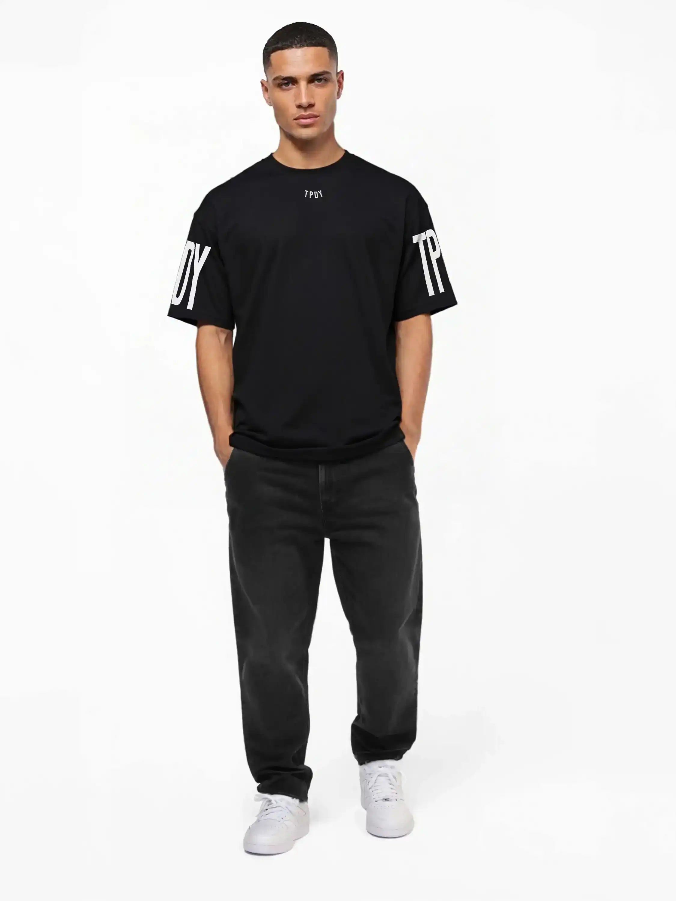 Ein Mann blickt mit den Händen in den Taschen nach vorne und trägt das trueprodigy Viktor Oversize-Herren-T-Shirt mit Front- und Ärmelprints, gepaart mit dunklen Jeans und weißen Turnschuhen vor einem schlichten weißen Hintergrund.