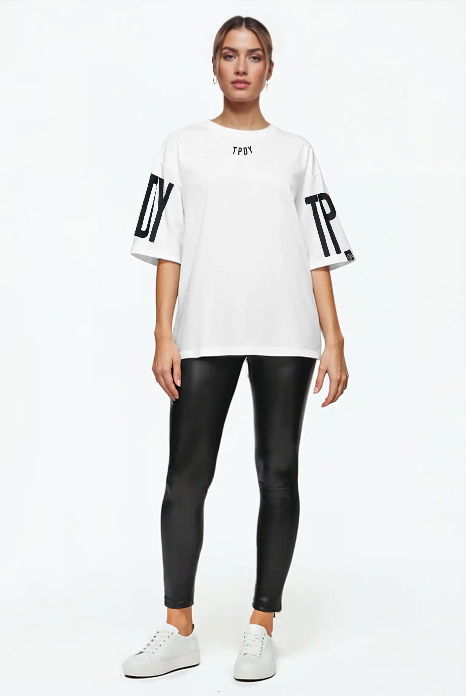 Eine Person blickt vor weißem Hintergrund nach vorne und trägt das trueprodigy Viktor F Oversize-Damen-T-Shirt mit Ärmelprints und Frontprint unter dem Kragen, gepaart mit einer schwarzen, taillierten Hose und weißen Sneakers.