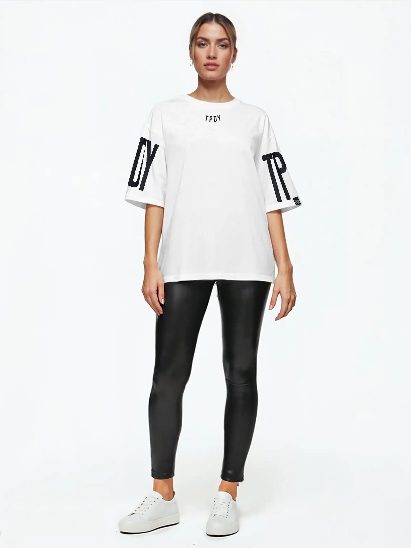 Eine Person blickt vor weißem Hintergrund nach vorne und trägt das trueprodigy Viktor F Oversize-Damen-T-Shirt mit Ärmelprints und Frontprint unter dem Kragen, gepaart mit einer schwarzen, taillierten Hose und weißen Sneakers.