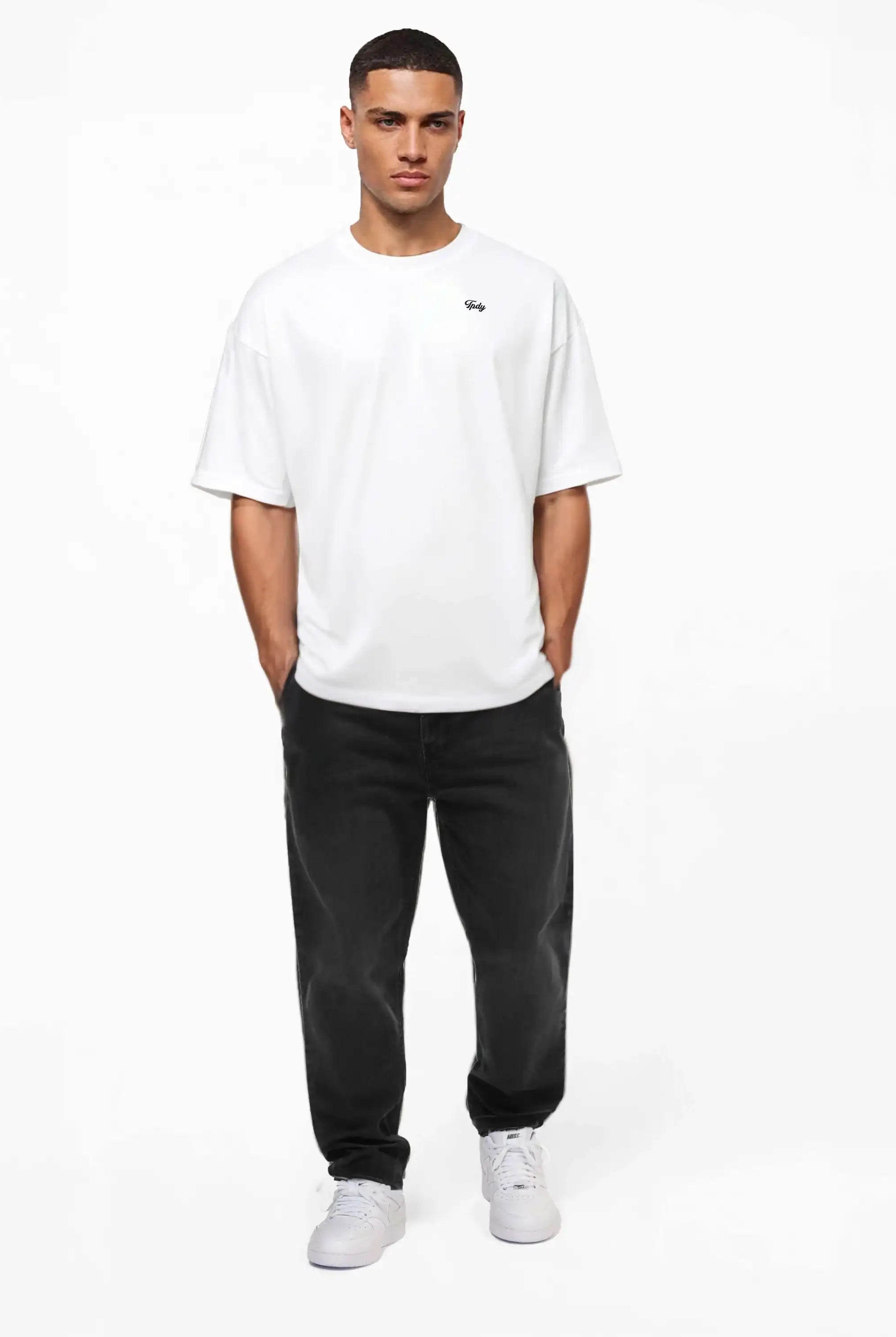 Ein Mann mit kurzen Haaren und den Händen in den Taschen steht vor einem weißen Hintergrund. Er trägt das trueprodigy Nika Oversize-Basicshirt mit TPDY-Stickerei & klarer Silhouette, schwarze Relaxed-Fit-Jeans und weiße Sneakers.