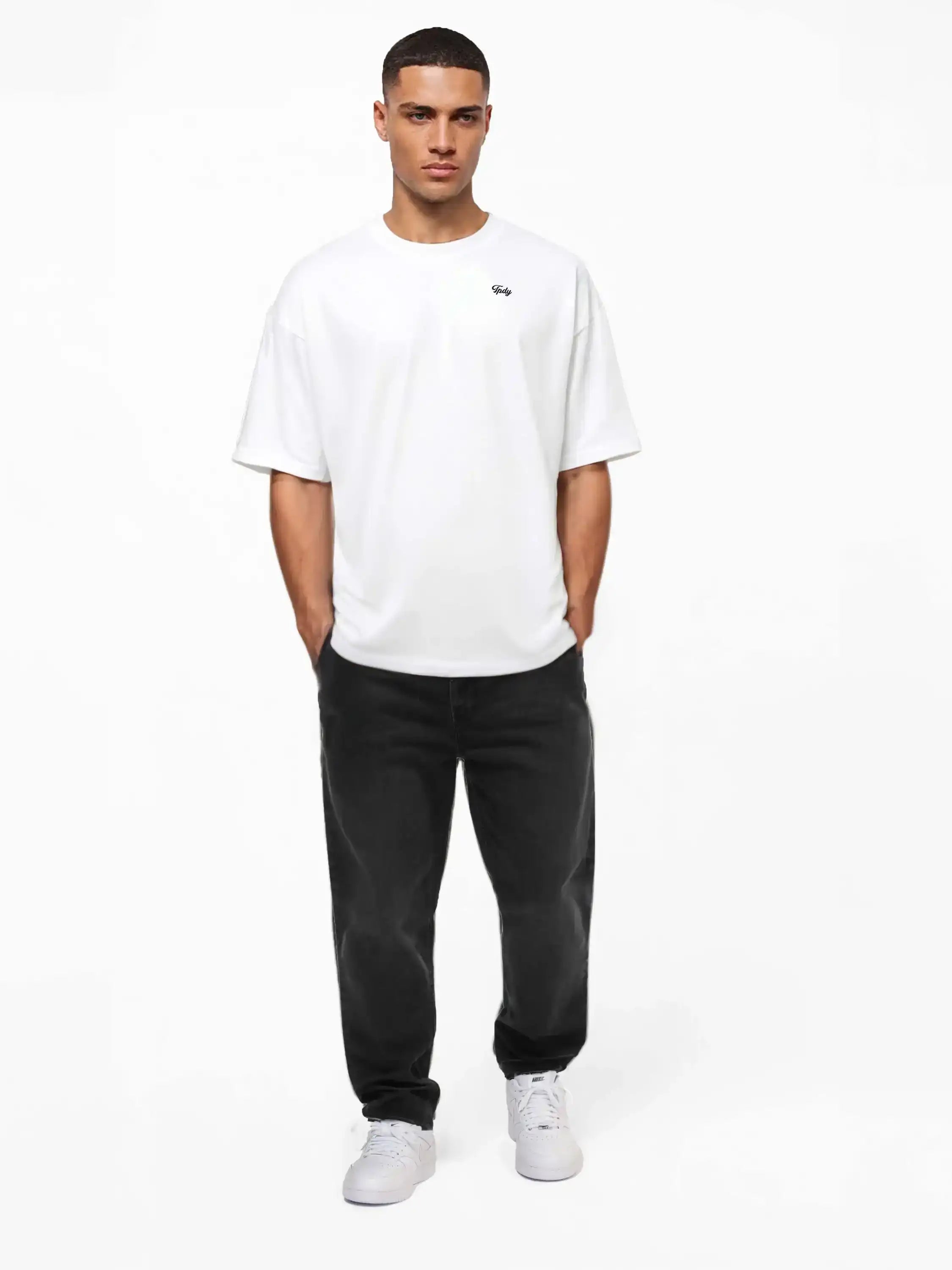 Ein Mann mit kurzen Haaren und den Händen in den Taschen steht vor einem weißen Hintergrund. Er trägt das trueprodigy Nika Oversize-Basicshirt mit TPDY-Stickerei & klarer Silhouette, schwarze Relaxed-Fit-Jeans und weiße Sneakers.