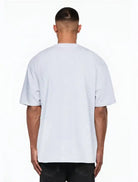 Ein Mann, der mit dem Rücken zur Kamera steht, trägt das trueprodigy Nika Oversize-Basicshirt mit TPDY-Stickerei & klarer Silhouette in hellgrau und schwarzer Hose, posiert vor einem weißen Hintergrund.