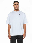 Ein Mann sieht nach vorne und trägt ein lockeres, hellgraues Nika Oversize-Basicshirt mit TPDY-Stickerei & klarer Silhouette von trueprodigy mit kurzen Ärmeln und einem kleinen Logo auf der linken Brust, gepaart mit einer schwarzen Hose. Der Hintergrund ist einfarbig weiß.