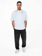 Ein Mann blickt nach vorne und trägt das trueprodigy Nika Oversize-Basicshirt mit TPDY-Stickerei & klarer Silhouette in Hellgrau, gepaart mit schwarzer Jeans und weißen Sneakers. Er steht vor einem schlichten weißen Hintergrund mit Händen in den Taschen und einem neutralen Gesichtsausdruck.