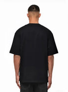 Eine Person mit kurzen Haaren schaut von der Kamera weg und trägt ein schwarzes trueprodigy Nika Oversize-Basicshirt mit TPDY-Stickerei & klarer Silhouette und dunkle Jeans vor weißem Hintergrund.