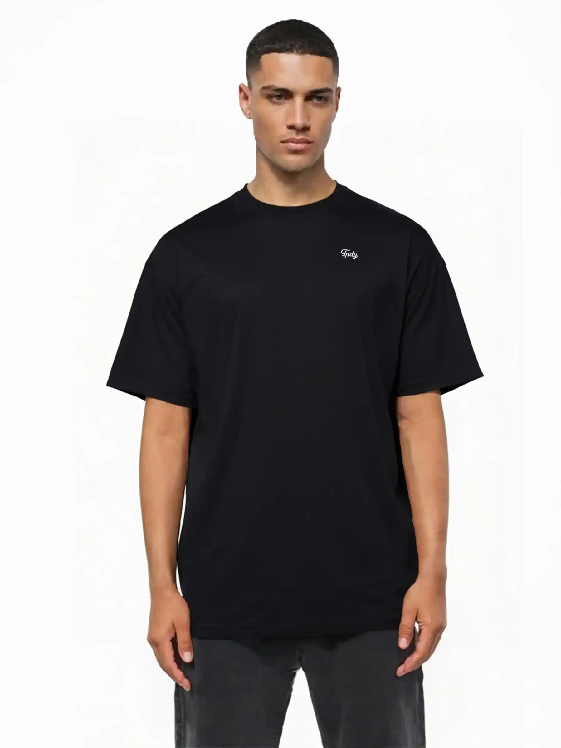 Ein Mann blickt nach vorne im Nika Oversize-Basicshirt mit TPDY-Stickerei & klarer Silhouette von trueprodigy, einem schlichten schwarzen Unisex-T-Shirt mit kleinem weißen Brustlogo und einer dunklen Jeans, die vor einem einfarbig weißen Hintergrund steht.