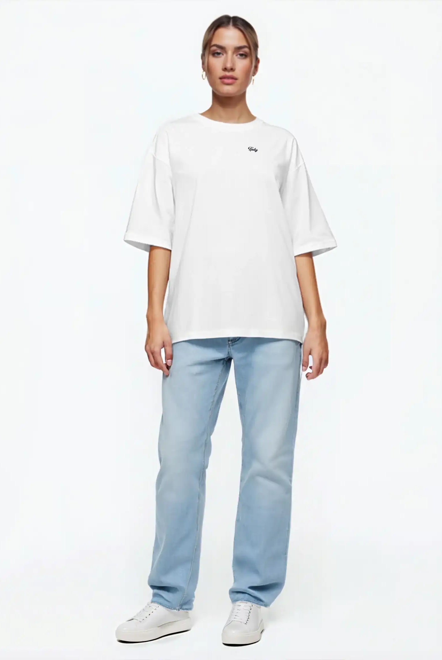 Eine Person steht mit dem Gesicht nach vorne vor einem schlichten weißen Hintergrund und trägt das trueprodigy Nika F Damen Oversize-T-Shirt mit TPDY-Stickerei auf der Brust, gepaart mit hellblauen Jeans und weißen Sneakers.