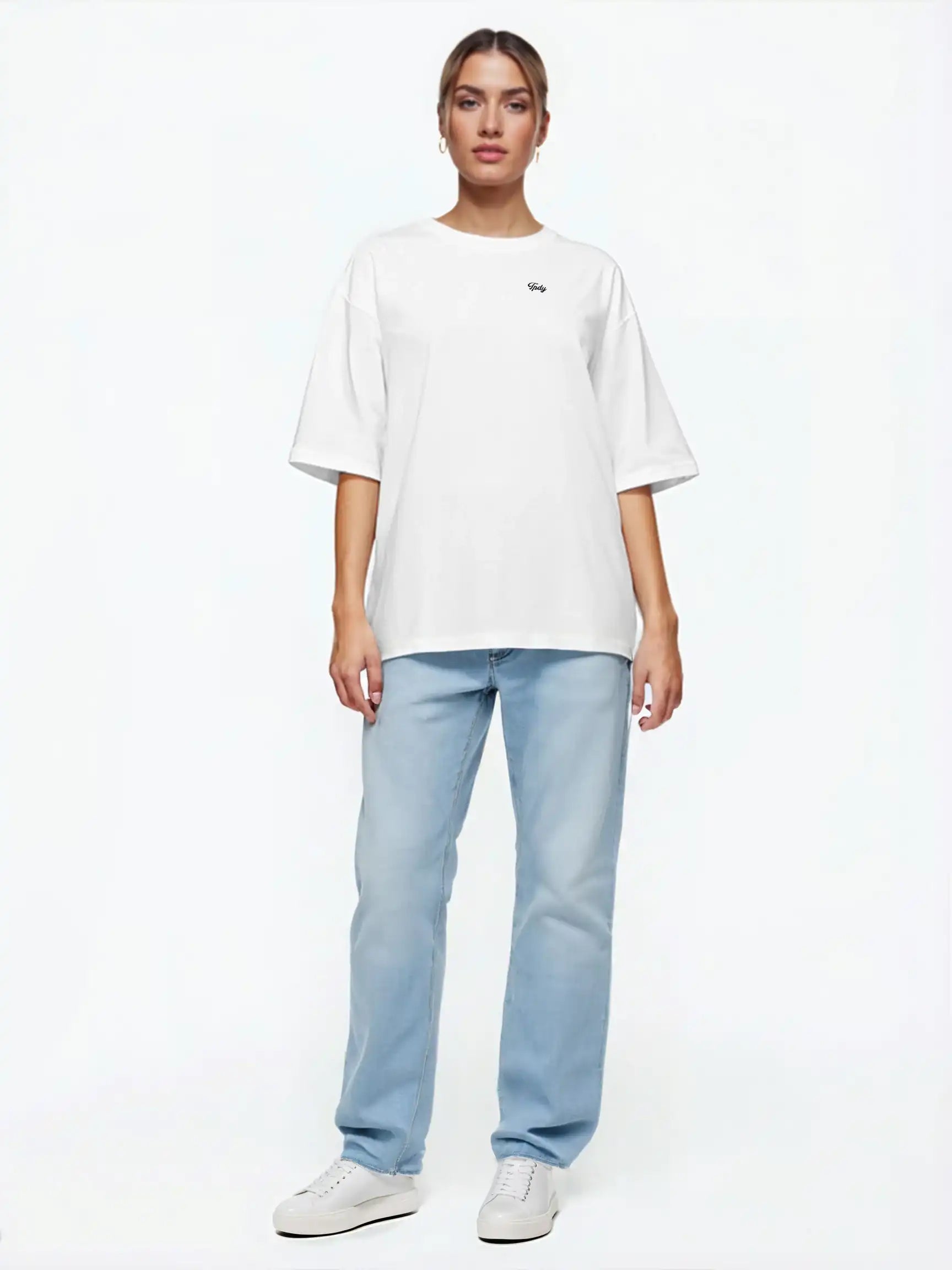 Eine Person steht mit dem Gesicht nach vorne vor einem schlichten weißen Hintergrund und trägt das trueprodigy Nika F Damen Oversize-T-Shirt mit TPDY-Stickerei auf der Brust, gepaart mit hellblauen Jeans und weißen Sneakers.