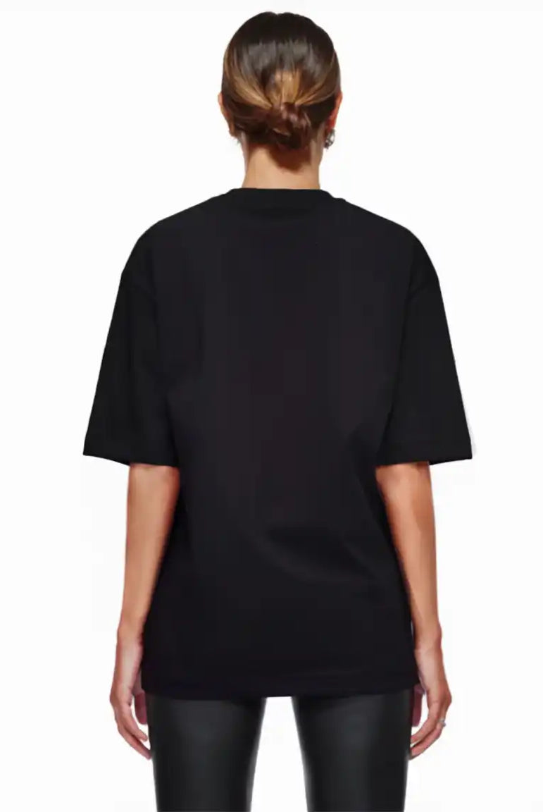 Eine Person mit braunen Haaren in einem tiefen Dutt wird von hinten gezeigt, die das trueprodigy Nika F Damen Oversize-T-Shirt mit TPDY-Stickerei auf der Brust und eine schwarze Hose vor einem schlichten weißen Hintergrund trägt.