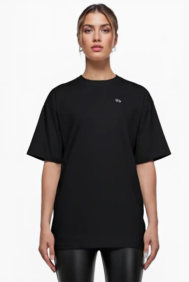 Eine Person sieht nach vorne und trägt das trueprodigy Nika F Damen Oversize-T-Shirt mit TPDY-Stickerei auf der Brust in schwarz mit kurzen Ärmeln, gepaart mit einer schwarzen Hose in Lederoptik. Der Hintergrund ist weiß.