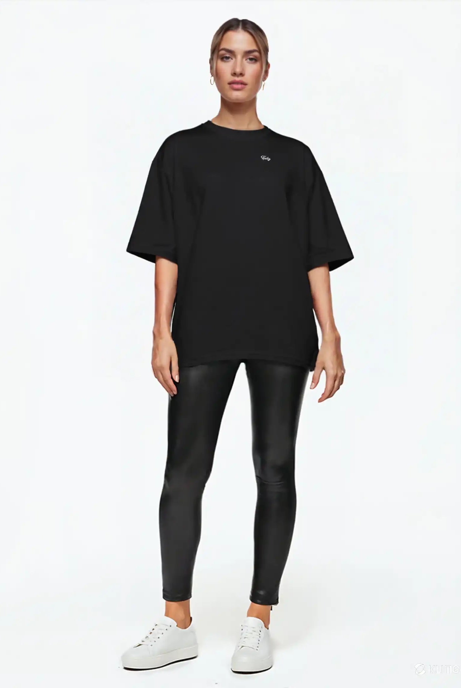 Eine Person steht mit dem Gesicht nach vorne vor einem schlichten weißen Hintergrund und trägt das trueprodigy Nika F Damen Oversize-T-Shirt mit TPDY-Stickerei auf der Brust, schwarz glänzende Leggings und weiße Turnschuhe. Die Haare sind zurückgebunden und die Arme liegen an der Seite.