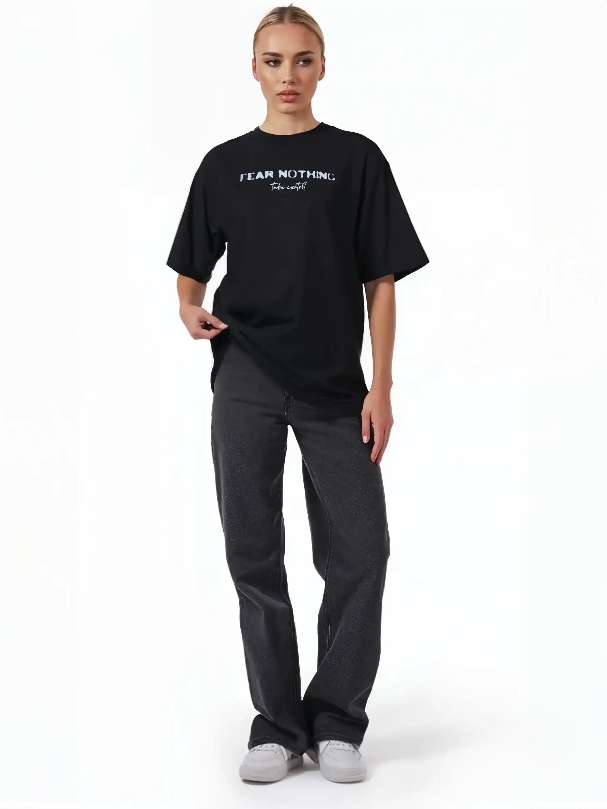 Eine Person steht mit dem trueprodigy Noa F Oversize-T-Shirt mit "FEAR NOTHING" & "take control"-Frontprint in schwarz und dunkelgrauer Jeans, mit hellen, zurückgebundenen Haaren und hellen Turnschuhen vor einem schlichten weißen Hintergrund.